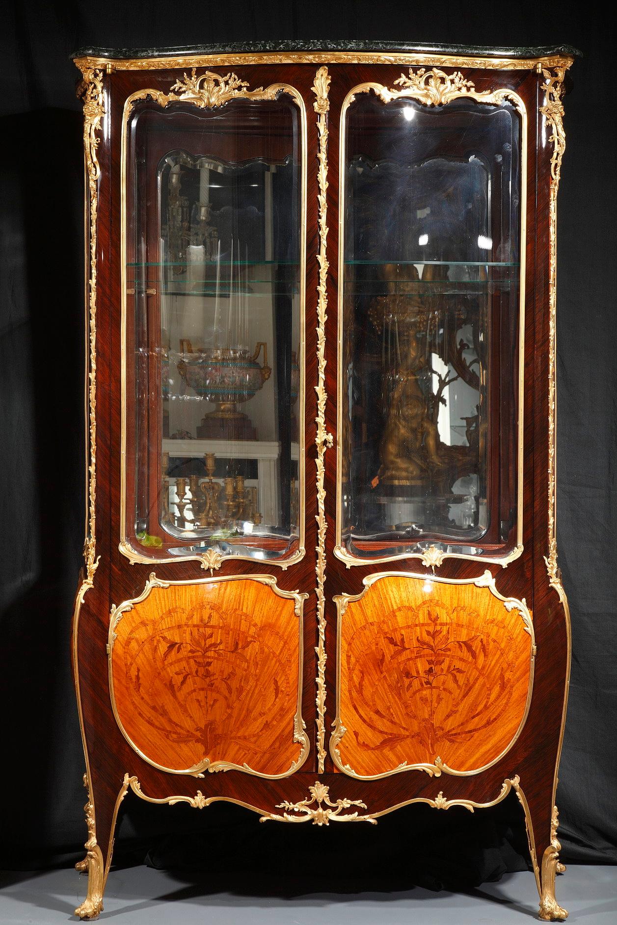 Elegant vitrine in veneered wood and gilded bronze. Opening with two curved doors with foliated frame in gilded and chiseled bronze, it is composed in the upper part of bevelled glass and in the lower part of « end-cut » floral inlays. Resting on
