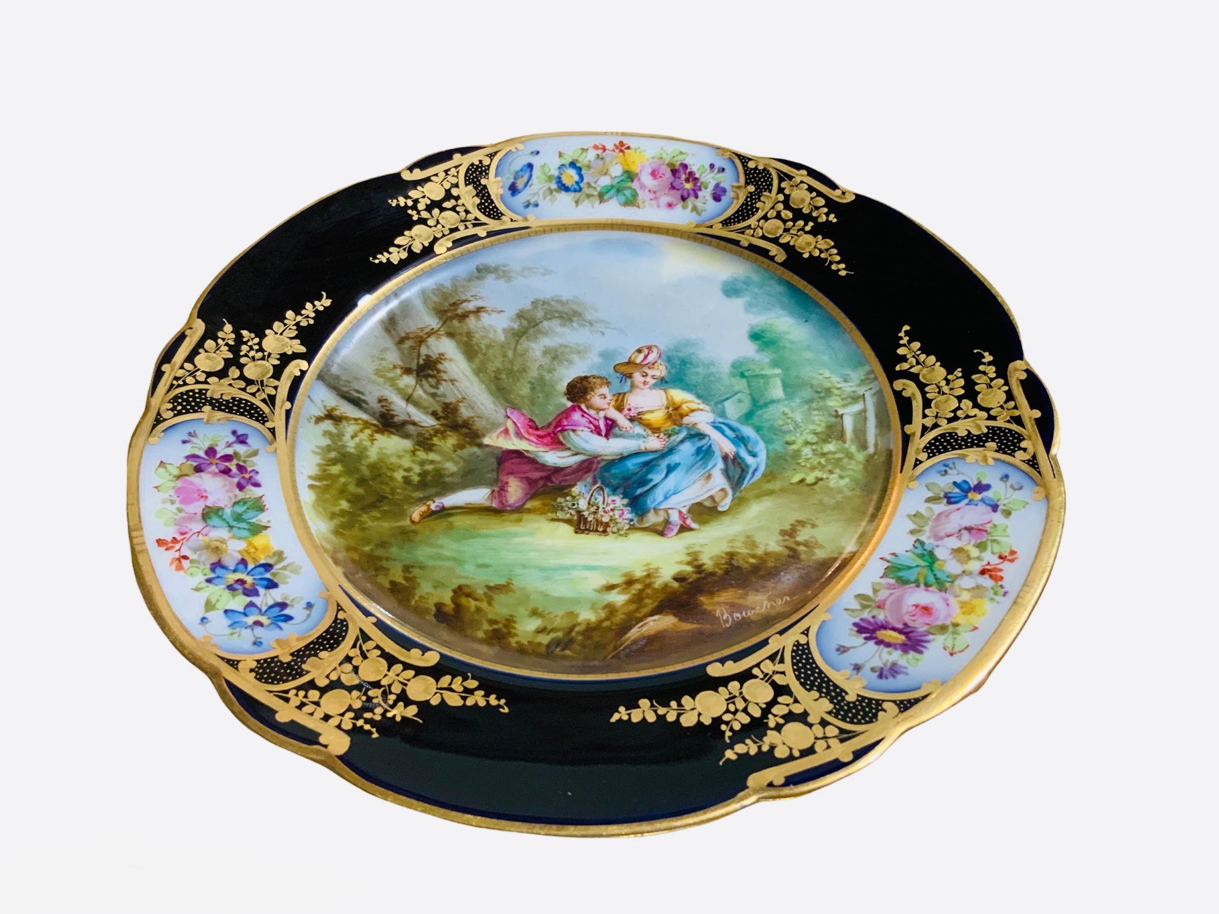 This is a Chateau de Tuileries Sevres Style hand painted cabinet porcelain plate. It depicts a pastoral scene of a courting couple in the center with a gilt cobalt blue circular rim. The rim is adorned with three bouquets of flowers framed with gilt