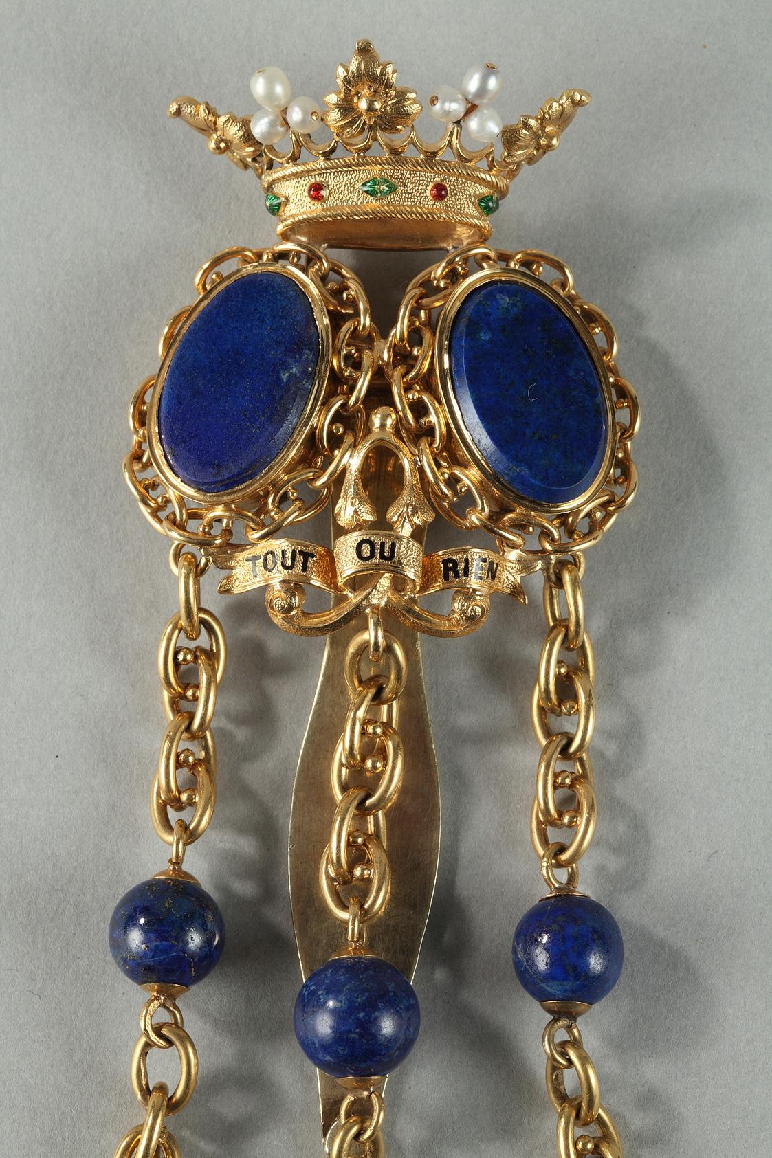 Gold chatelaine with belt hook behind. Two smooth lapis lazuli blazons are set in think, chain-link rings and topped with a crown embellished with pearls and inlaid with small green and red stones. Below the lapis lazuli blazons is a black enamel