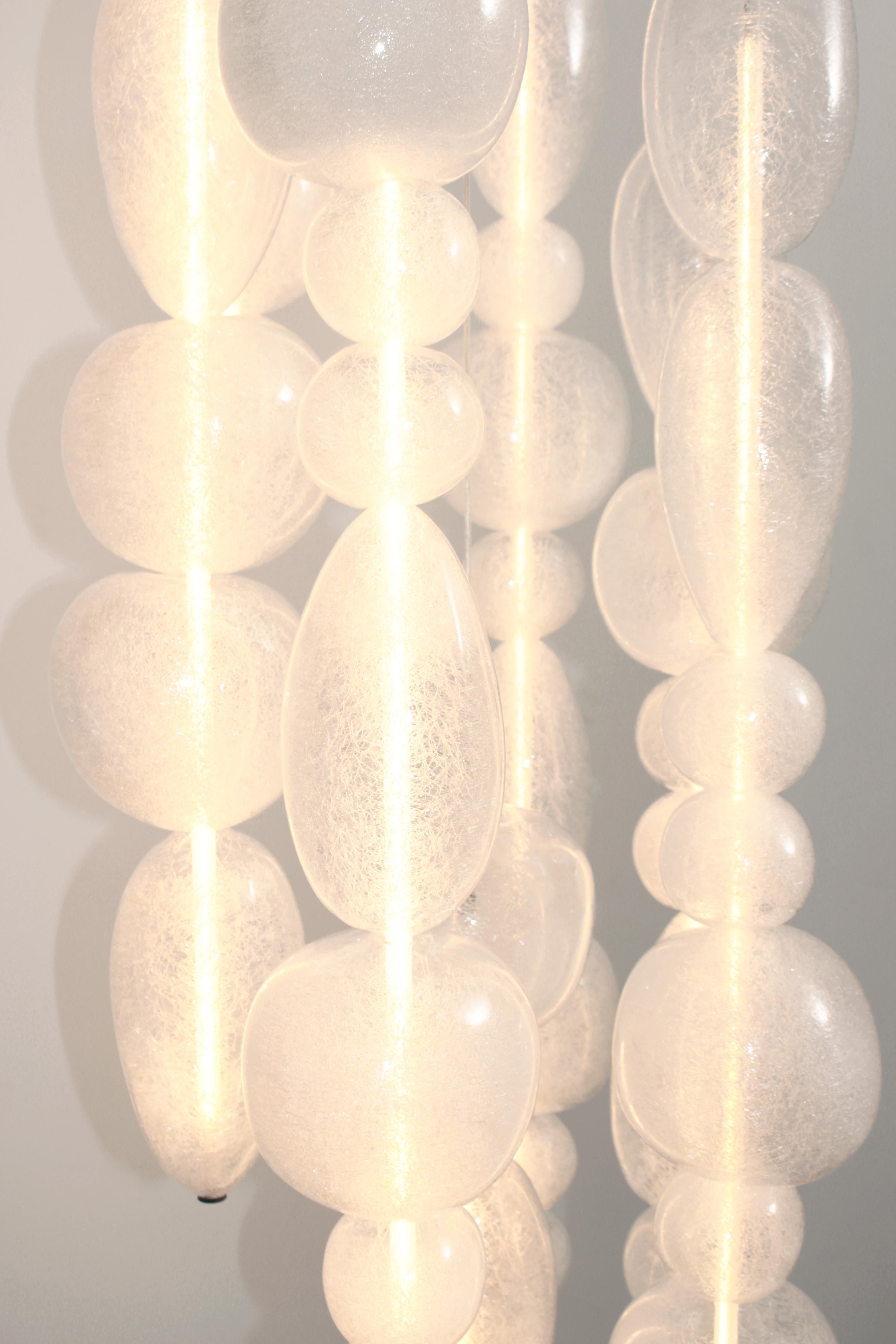 Mouth-blown transparent glass pendants. 4 glass columns on 42 cm round canopy.

The Chavana collection features pebble inspired glasses stacked along a 17W LED tube, lighting up to form a cascade of light and color. 
The smooth pebbles are filled