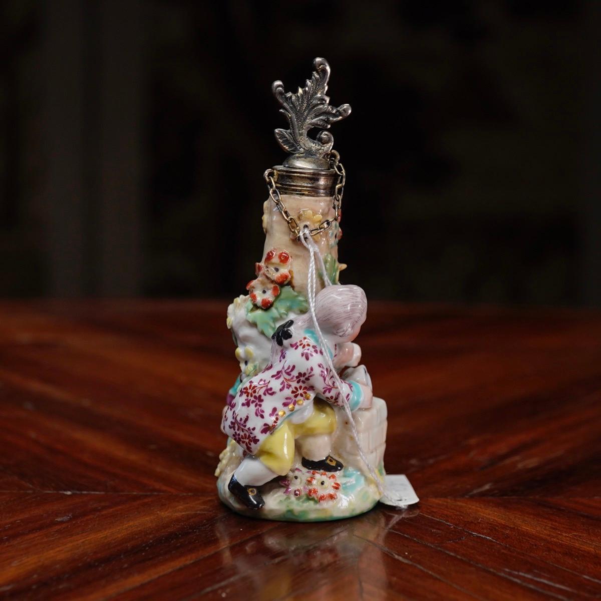 Chelsea scent bottle in the form of children building a house of cards against a tree trunk studded with flowers and leaves, the base with a gilt flower sprig within a turquoise dentil border, the neck with a silver mount with rococo leaf