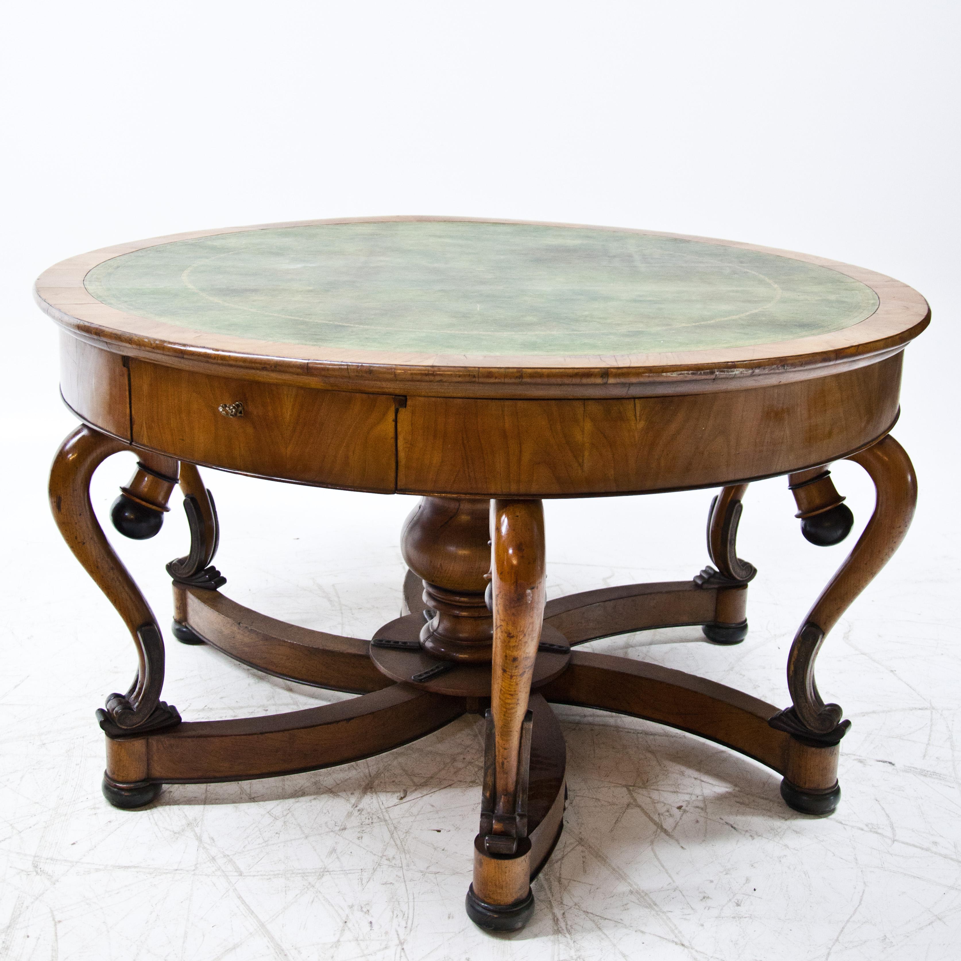 Round library table of cherrywood standing on six s-shaped legs with volute decoration. The struts are grouped radially around the balustered central foot. The high apron with two drawers ends with a slight profile edge, the table top is covered