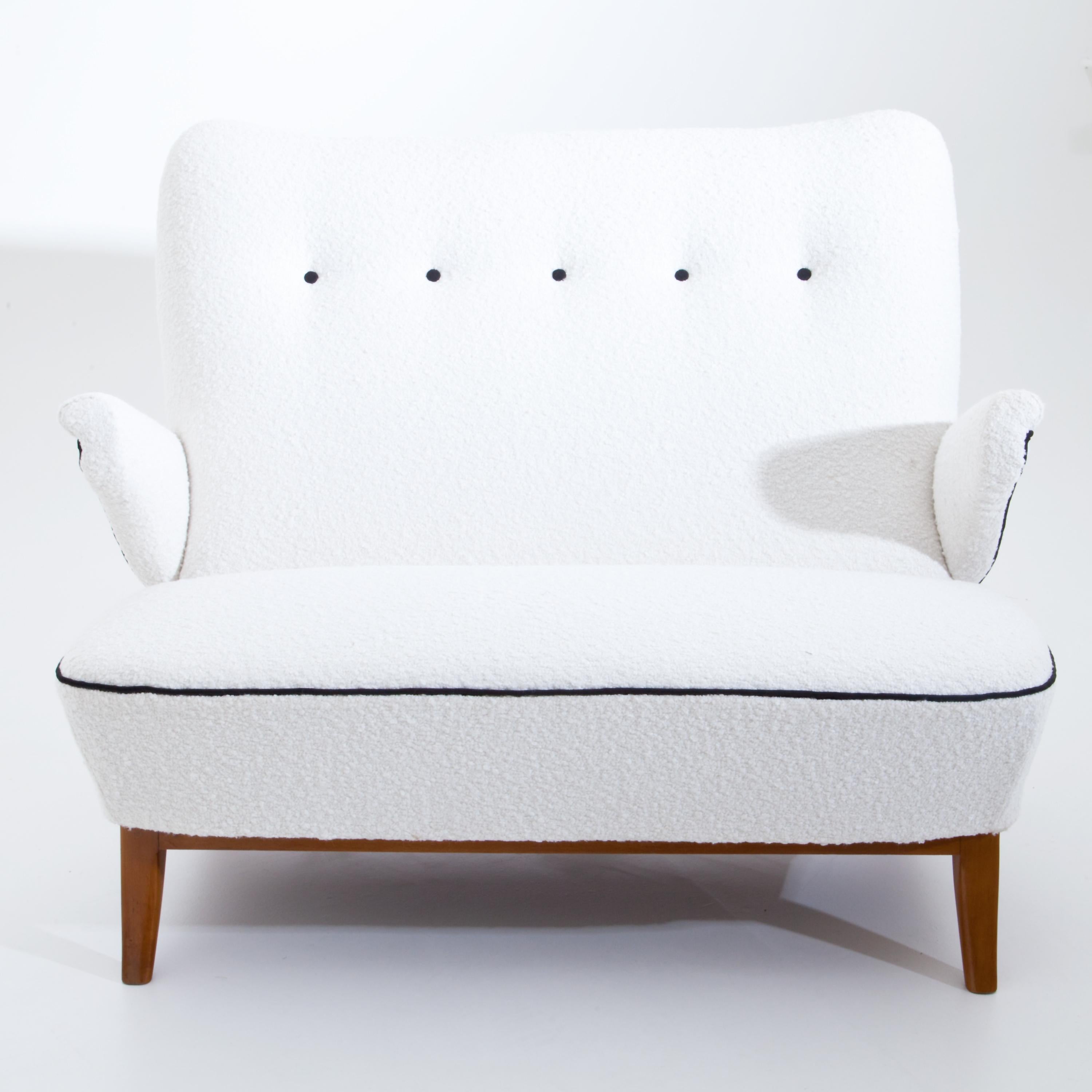Midcentury sofa or love seat on curved wooden feet. The strongly inclined, rounded backrest and the seat and armrests are newly covered with a white bouclé fabric with black piping and buttons. The backrest can be tilted in two stages by simply