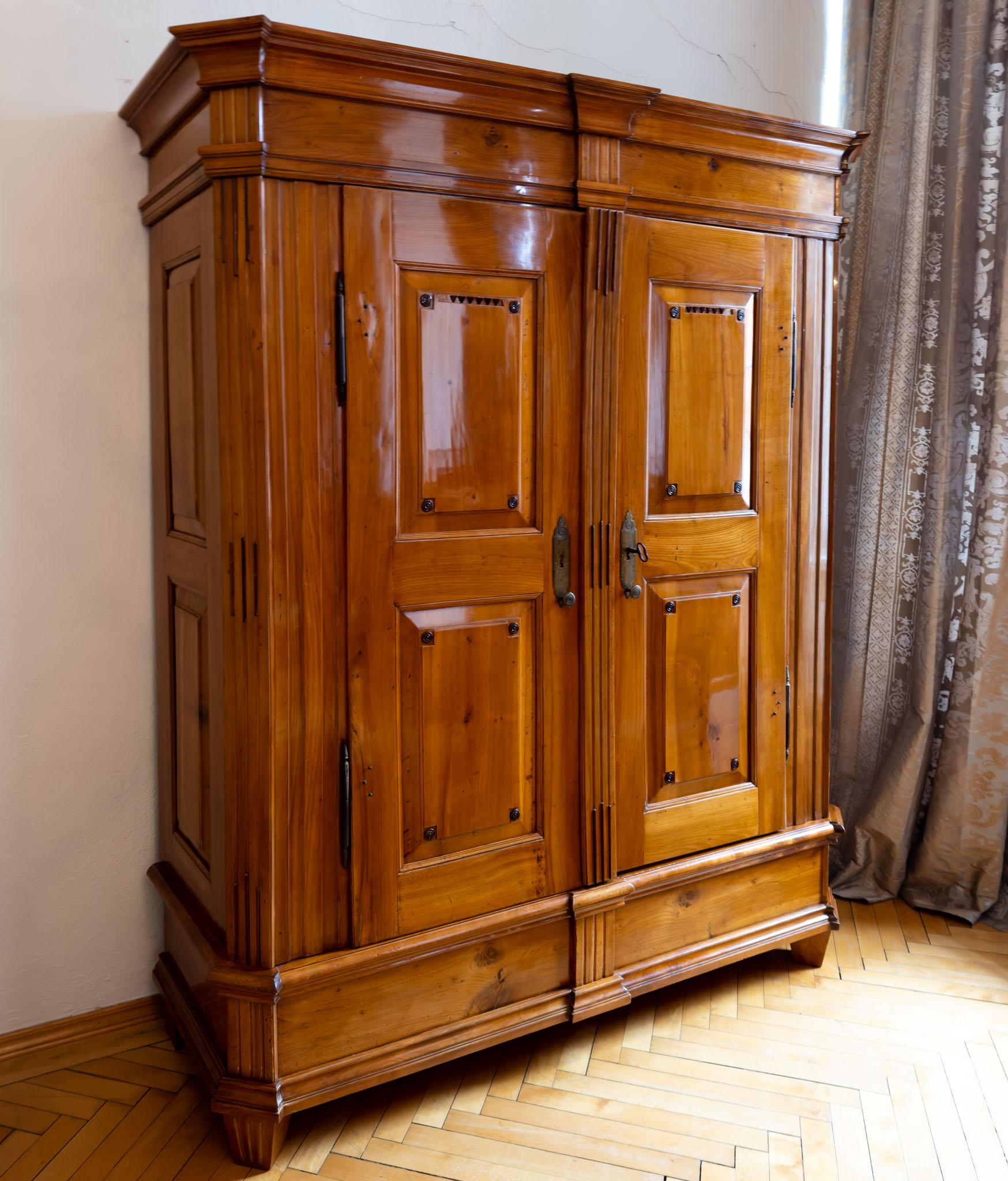 Two-door cabinet of cherry wood with filling fields, bevelled corners and fluted pilasters with pipe decoration. The corners of the panels are decorated with dark patinated discs. The high plinth and the cornice are profiled several times.