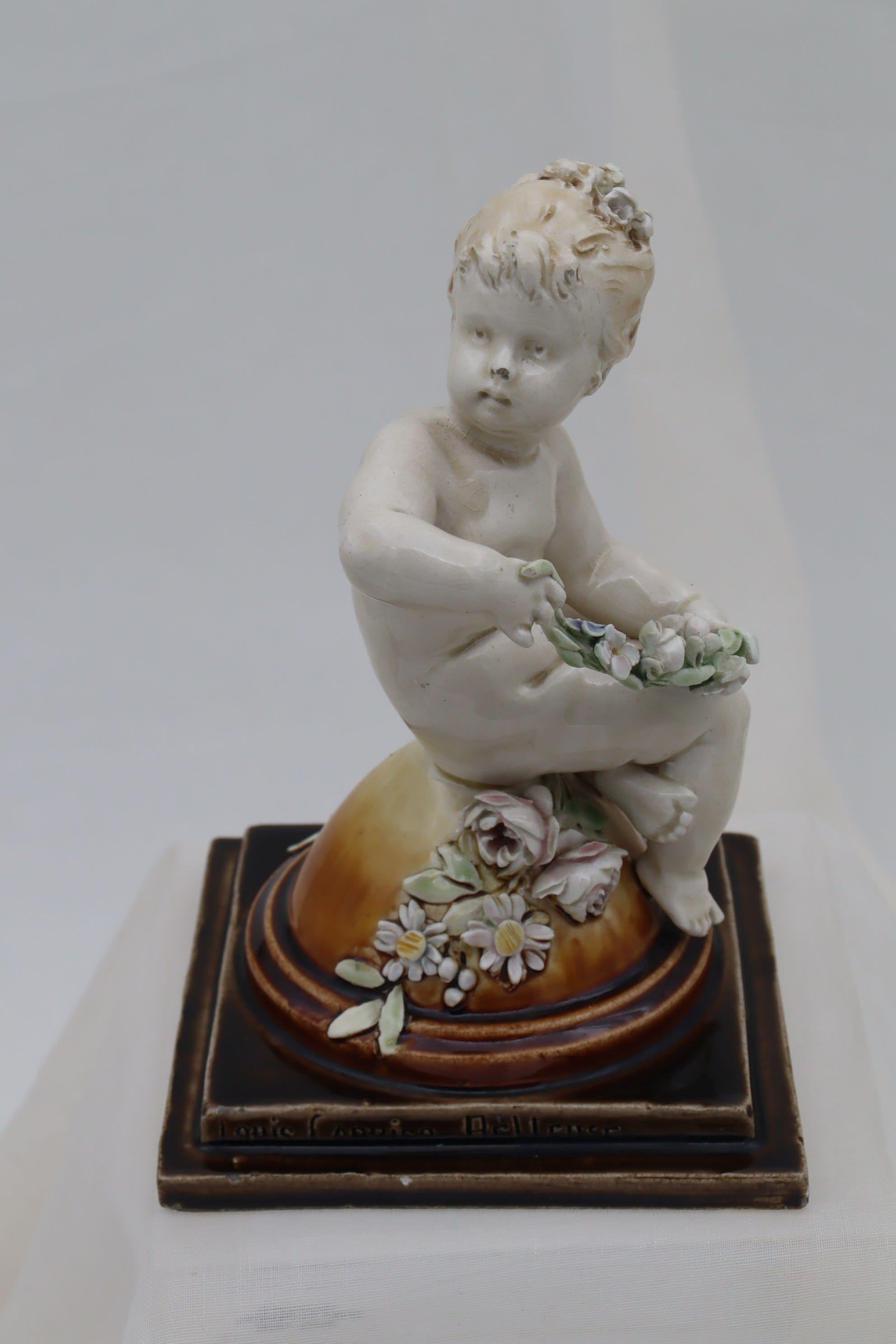 This cherub figurine was modelled by Louis Carrier-Belleuse (1848-1913) for the Choisy du Roi factory. The cherub is seated cross-legged on a dome, and holds a swag of flowers while another spray of flowers rests on the dome beneath her. The dome