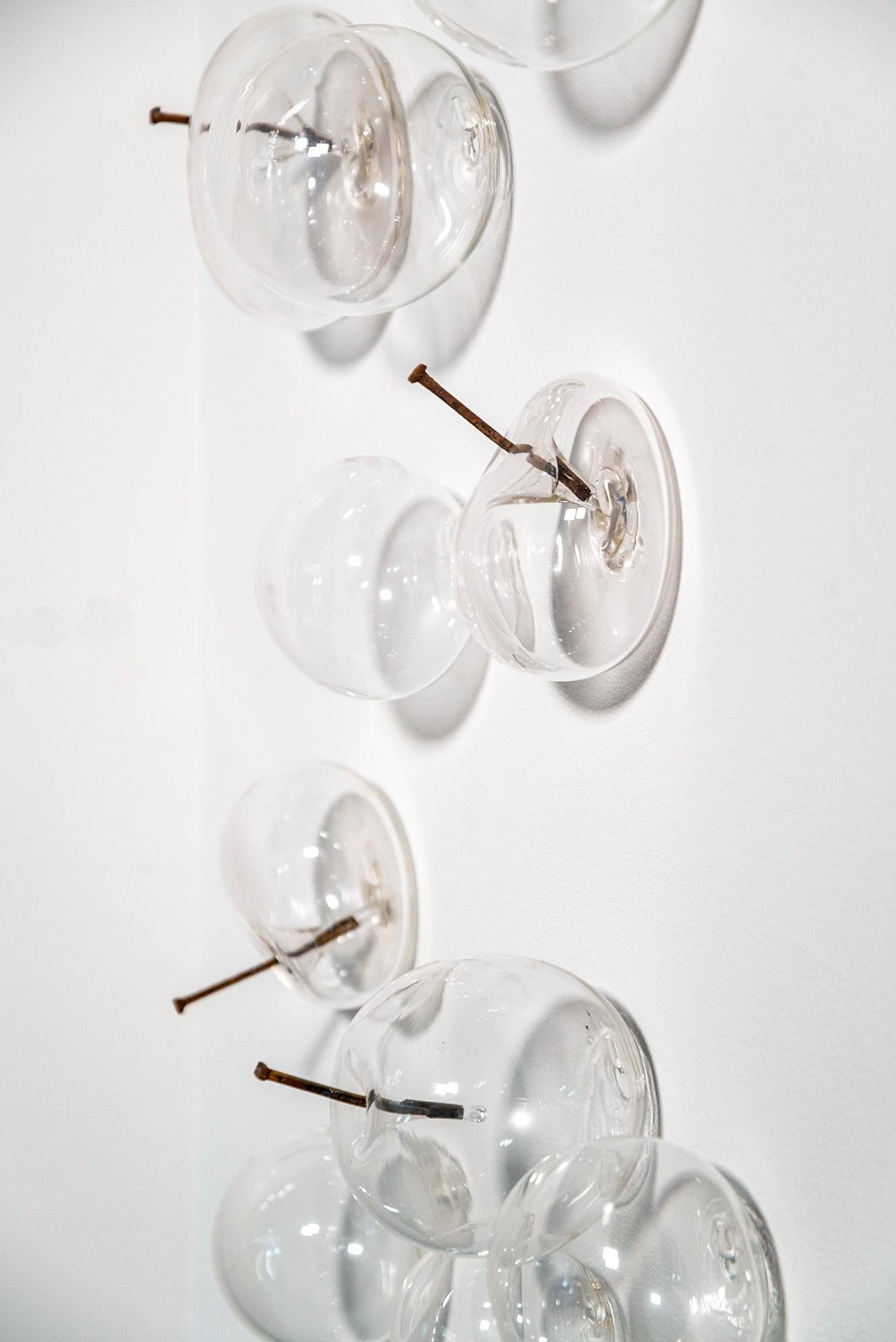 Cette sculpture murale contemporaine en verre a été créée par Cheryl Wilson Smith. Quatorze bulles en verre clair soufflé, certaines pleines, d'autres dégonflées, et percées de clous en fer créent une composition dynamique. La lumière traverse