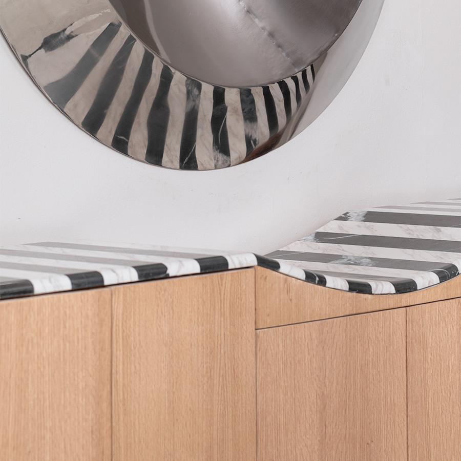 Inspired by the enigmatic smile of the Cheshire Cat, this sculptural credenza explores illusion, contrast, and refined craftsmanship.
Its top surface is composed of alternating strips of black and white marble, creating a mesmerizing rhythm that
