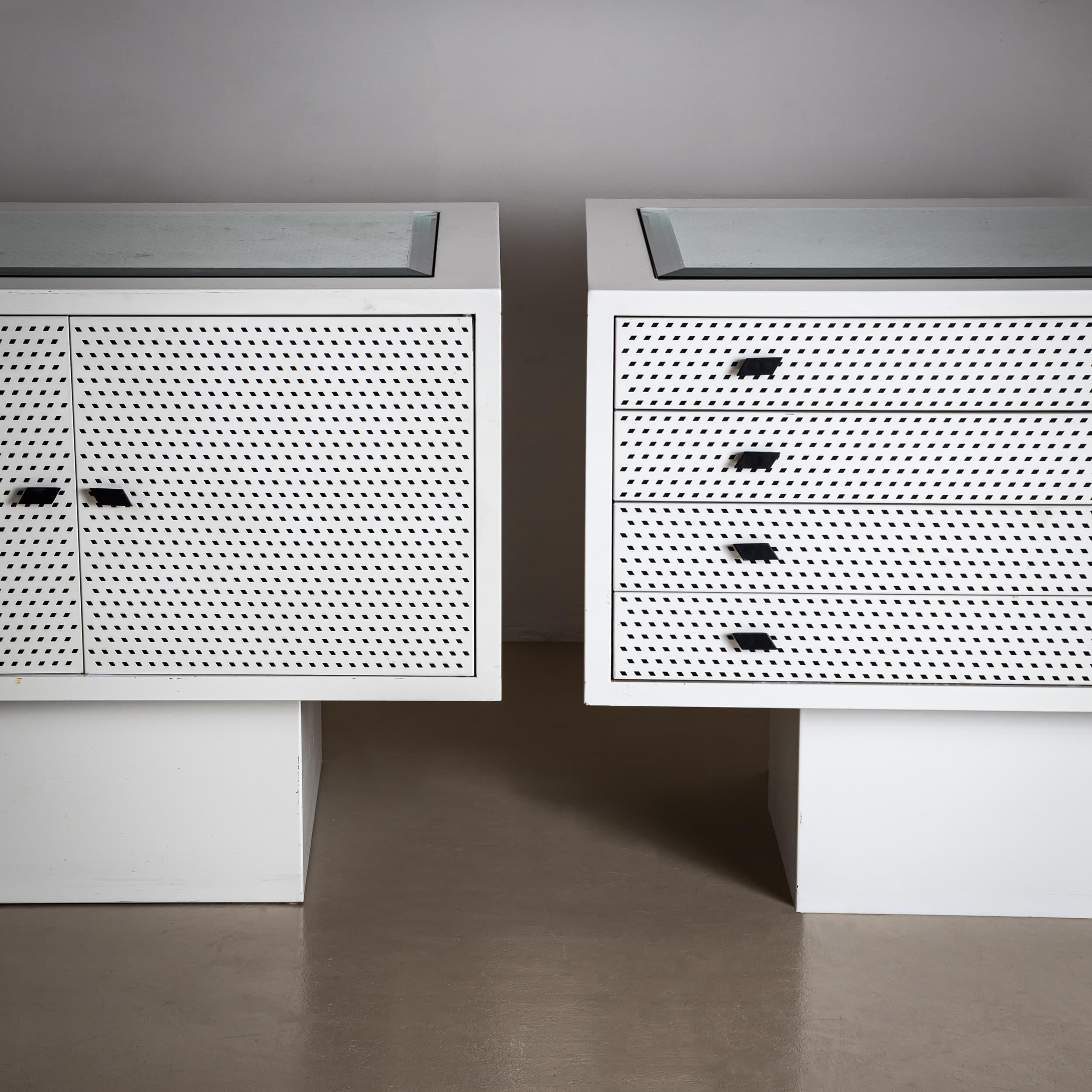 Comodino bianco con quattro cassetti e credenza a due ante su base quadrata, disegnato da Matteo Thun. I pannelli anteriori presentano un motivo traforato di piccoli rombi, ripresi nelle maniglie nere. Un pannello di vetro sfaccettato è incastonato