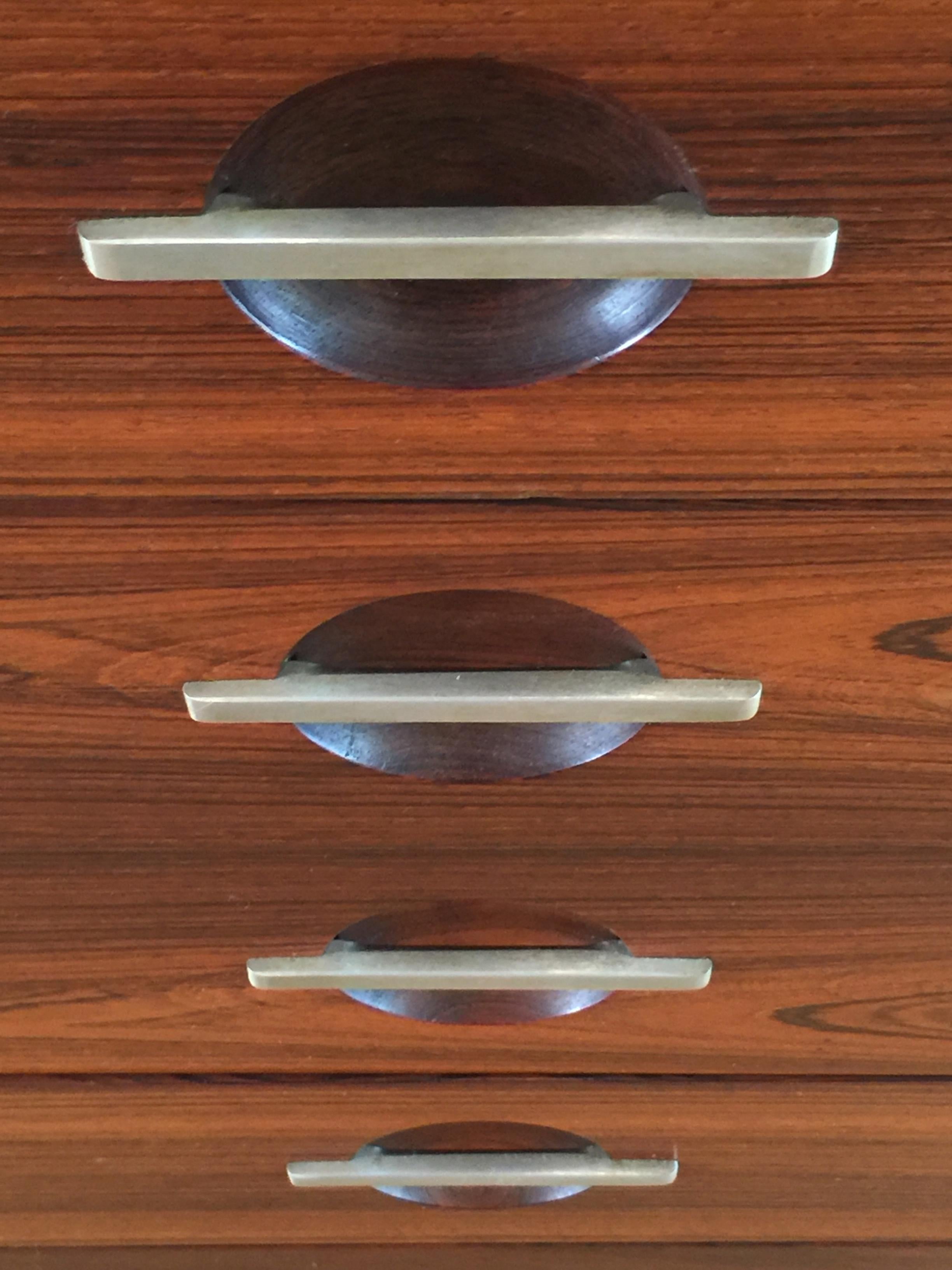 An exceptionally handsome chest of drawers in polished palisander contrasting with brushed stainless steel mounts. The drawer pulls are brushed steel across recessed hemispheres, and the stainless steel legs terminate in palisander 'feet'.

Illus.