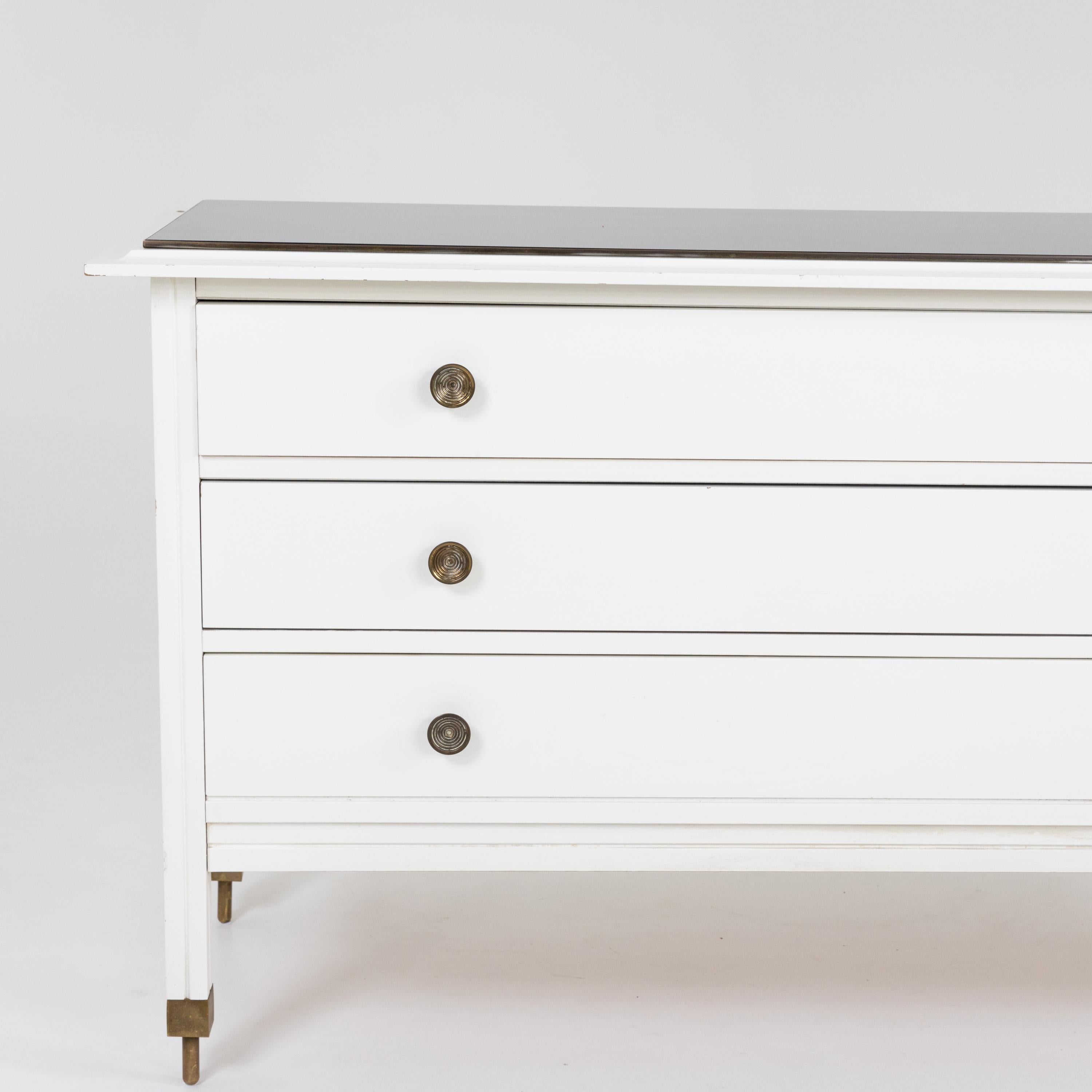 Carlo de Carli dresser with three drawers, standing on brass legs. The chest of drawers is painted in white and accented by round brass knobs and a black glass top.
