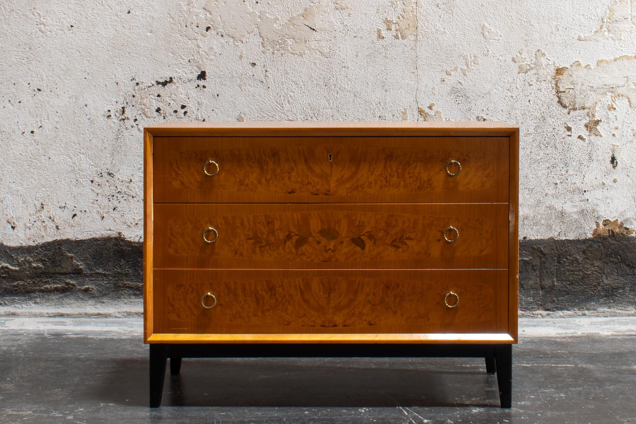 Art Deco chest of drawers in golden flame birch, featuring an ebonized base and intarsia inlay in a floral motif. Three drawers with brass pulls.