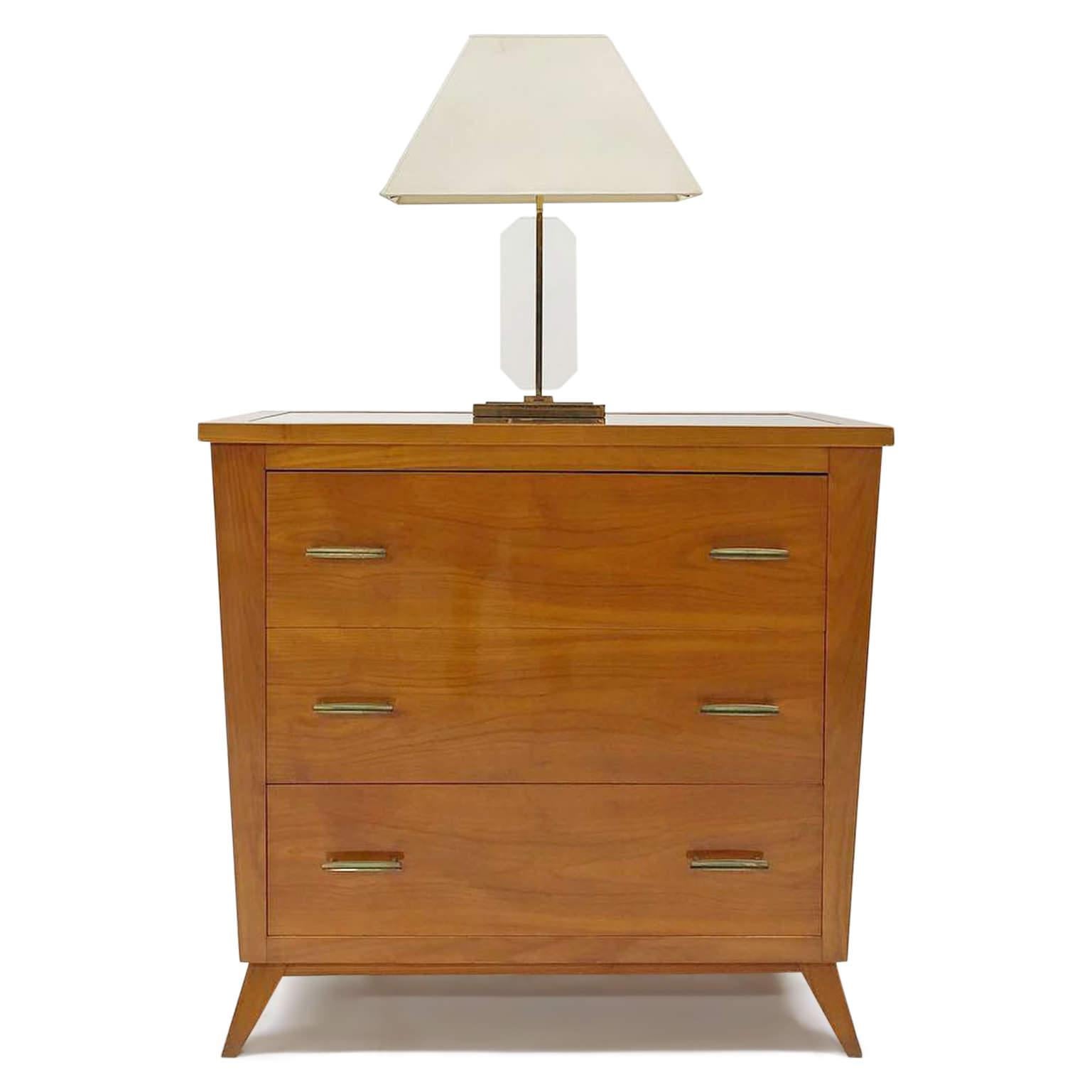 Three-drawered chest of drawers with a trapezoidal body and an imbedded glass top as well as brass handles. The chest is veneered with cherrywood.