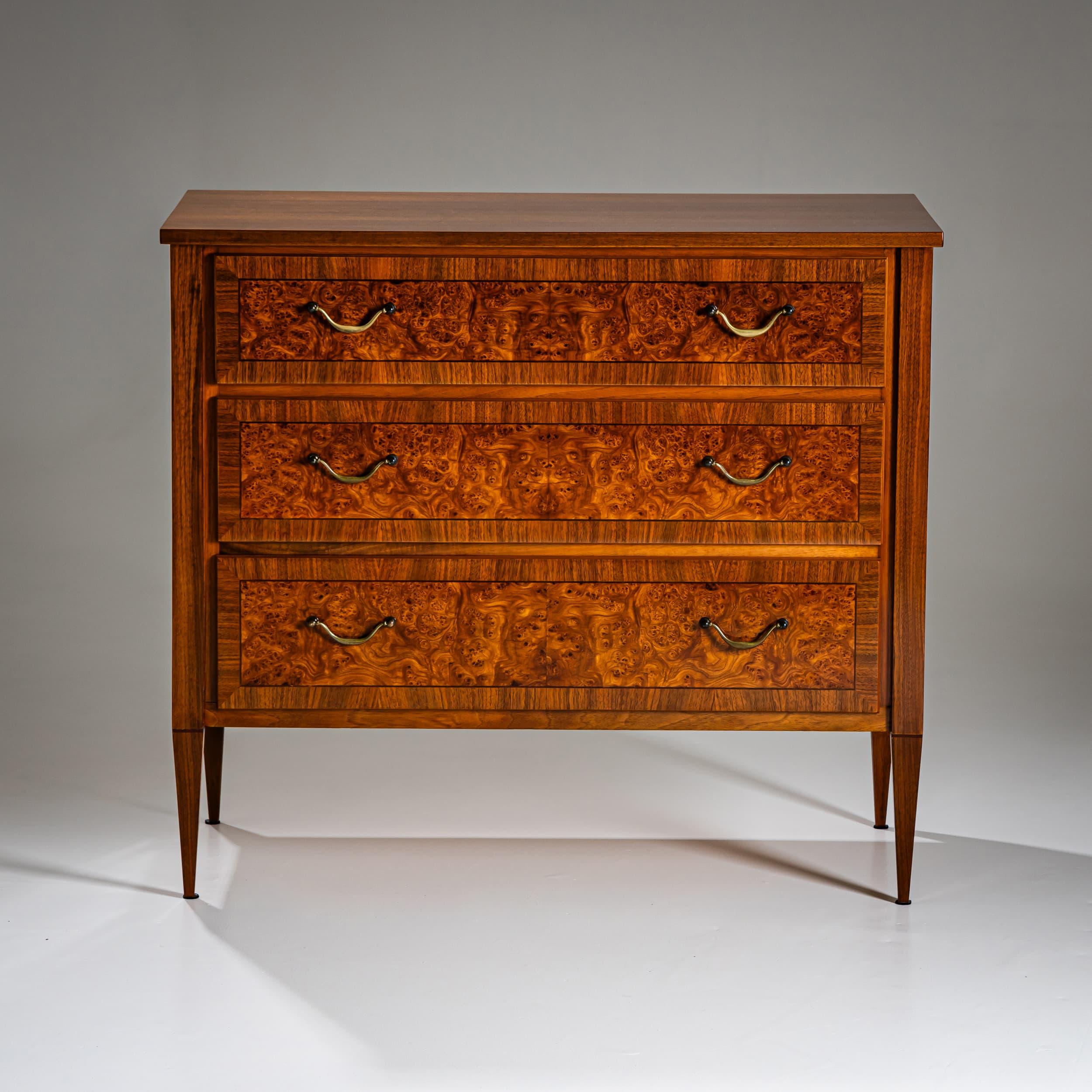 Small commode with three drawers and refined brass fittings. The piece stands on tapered square legs. The drawer fronts are bordered with framing elements and veneered in the center with richly grained burl wood.
