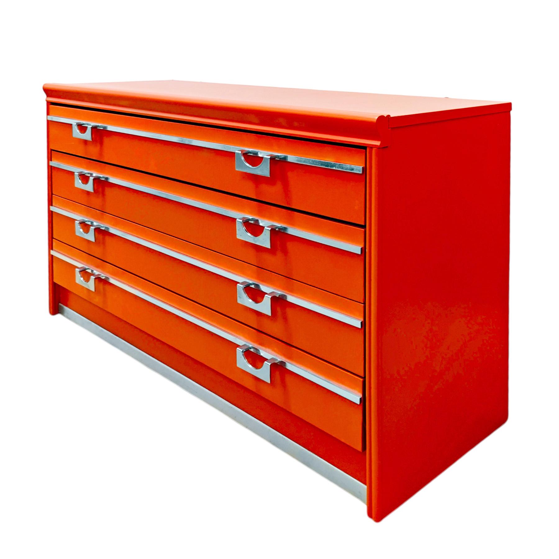 Divertida cómoda de madera maciza lacada en naranja y madera dura, con tiradores y adornos de metal niquelado.

España, 1970.

Dimensiones 
cm 100 x 37 + 2 (asas) x 62,5
pulgadas 39,37 x 14,57 + 0,79 (asas) x 24,60