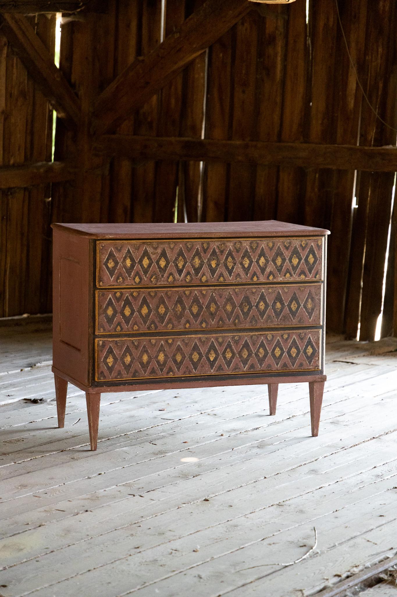 Brick-red chest of drawers with three drawers and coffered sides. The chest of drawers stands on square pointed feet and has been newly painted according to historical models. The front of the chest of drawers is embellished with a decorative