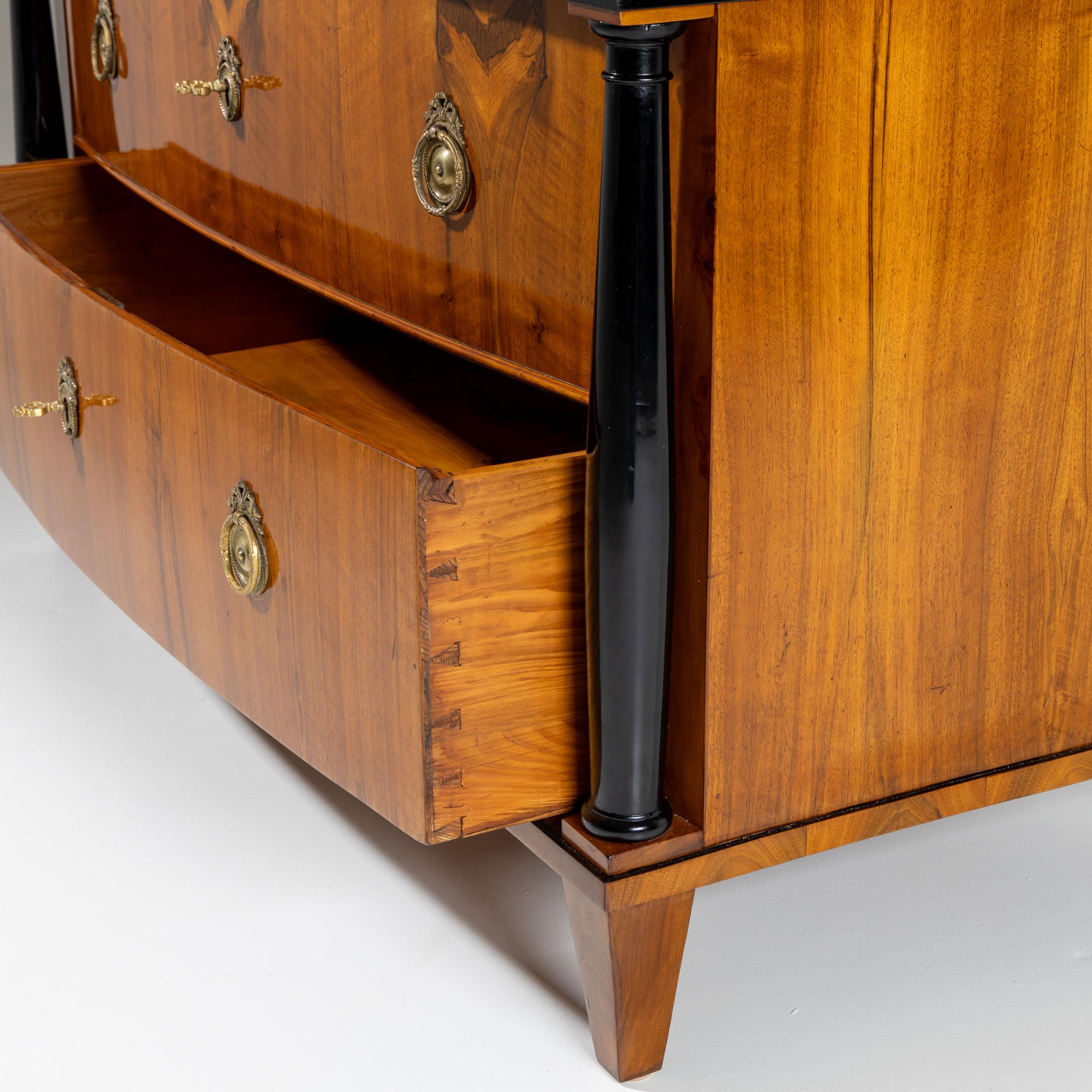 Three-drawer Biedermeier chest with a gently curved front and flanking ebonized full columns. Veneered in cherrywood and adorned with disc-shaped bronze mounts. The top drawer features three medallions depicting figural scenes created using the