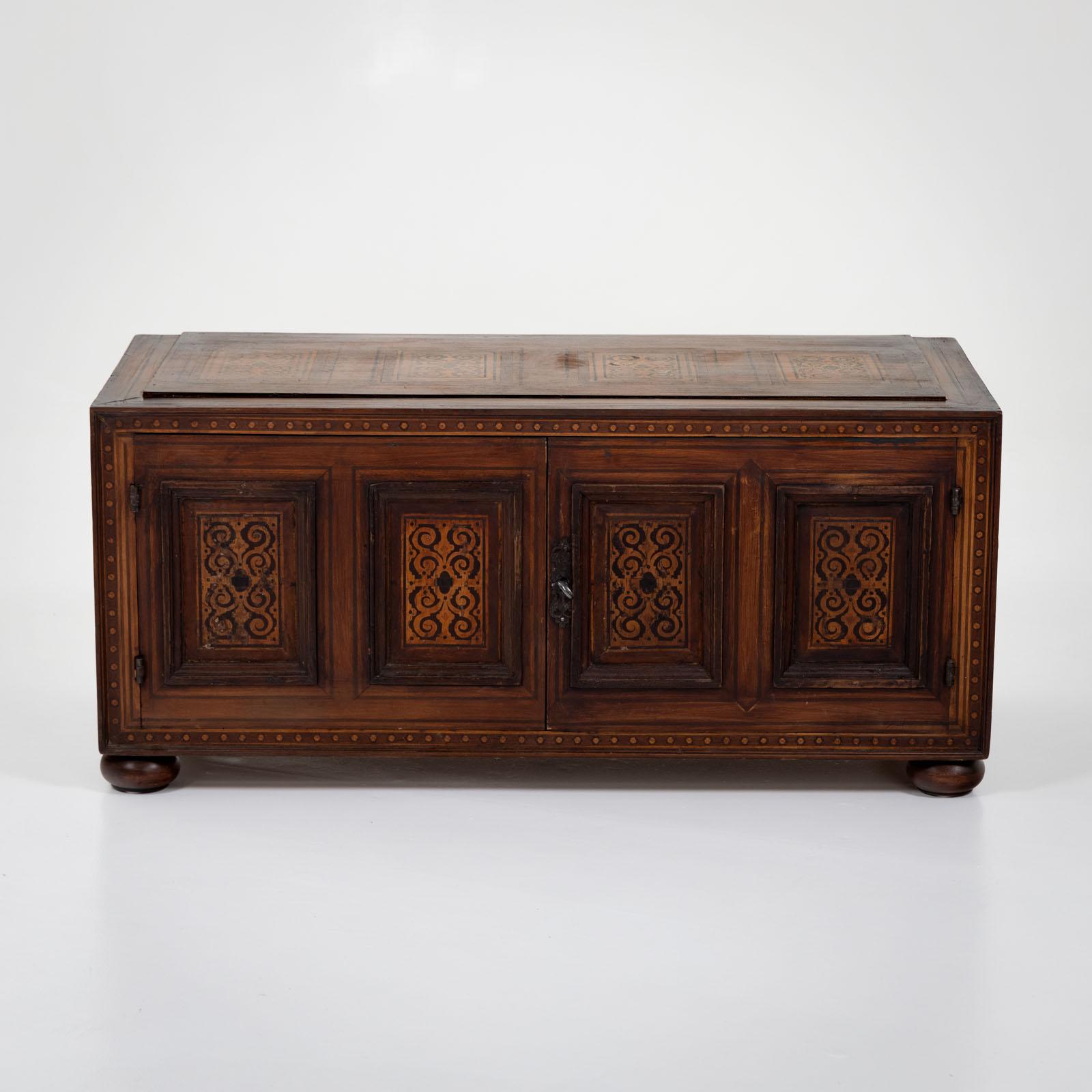 Chest with two doors and four panels with inlays and iron bands as well as handles on the sides. The chest stands on pressed ball feet and is fitted with a shelf inside. 