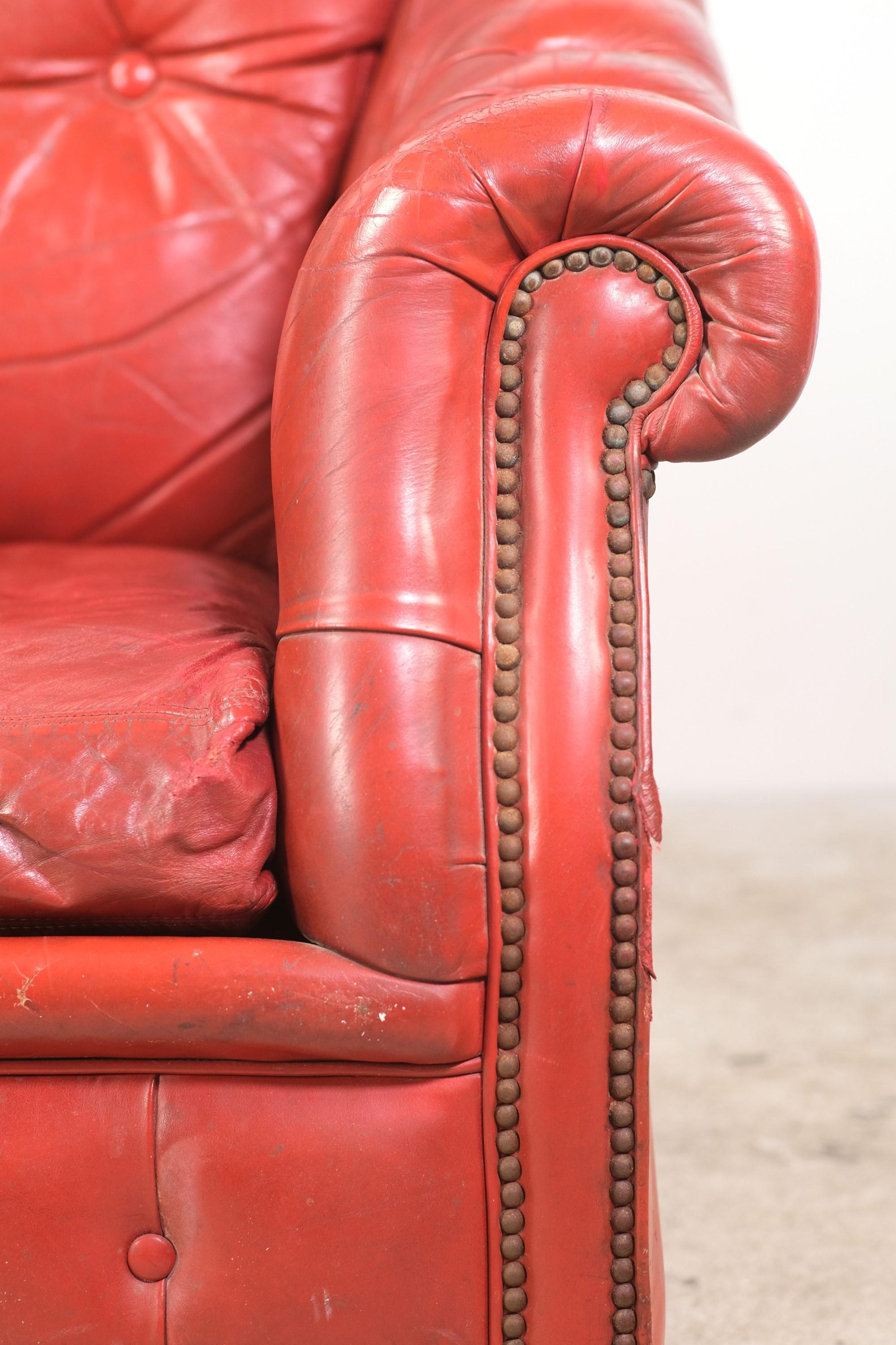 This European Chesterfield red armchair is tufted featuring deep buttons and studding along the trim. The front has round wooden feet and rolled arms with the back having red upholstered feet. This can be seen at our 400 Gilligan St location in