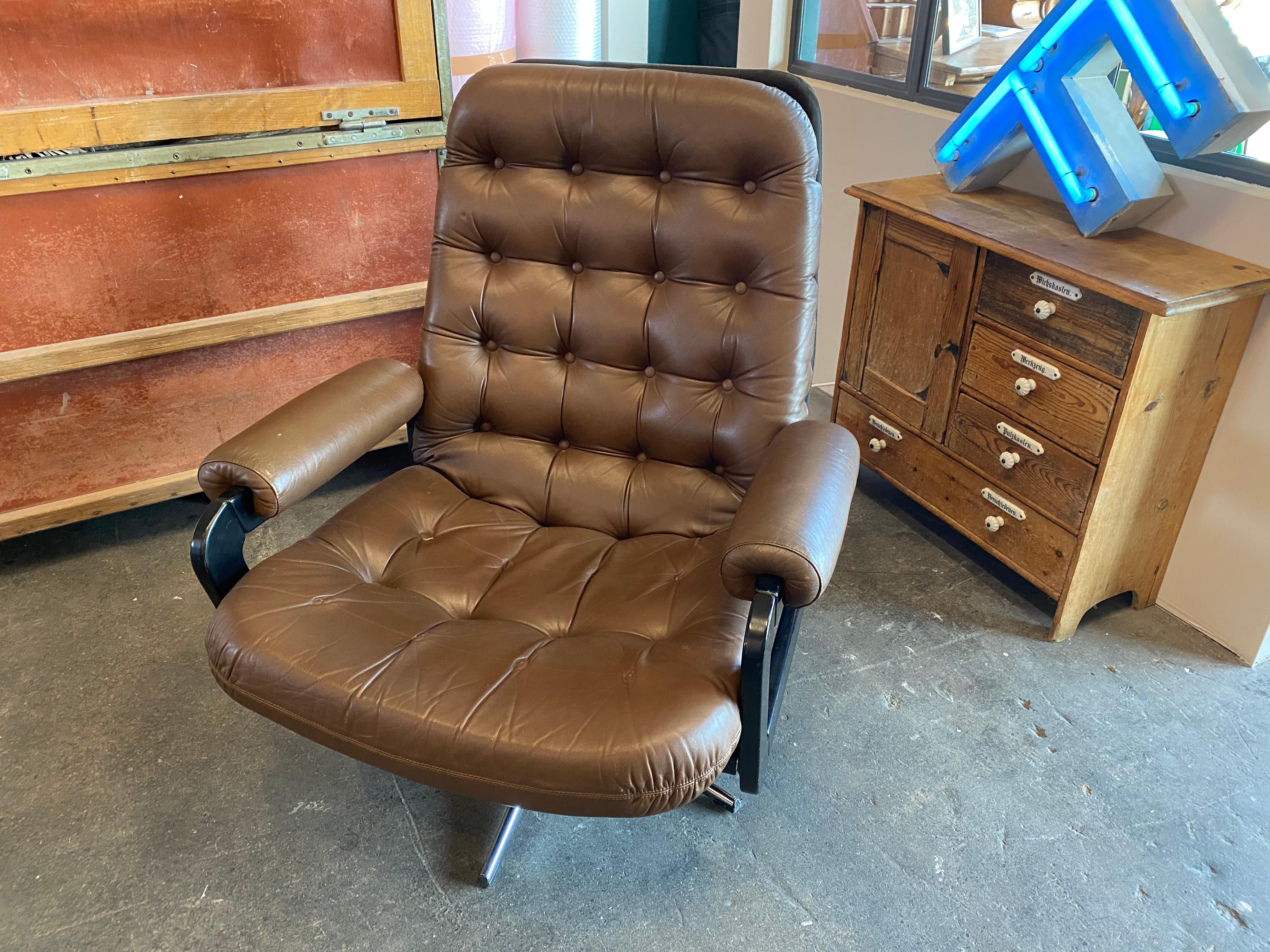 Chesterfield look lounge armchair designed in the 1970s. The armchair is upholstered, covered with brown faux leather and decorated with matching buttons. The base is chrome-plated. The design with the high back and armrests makes the armchair a