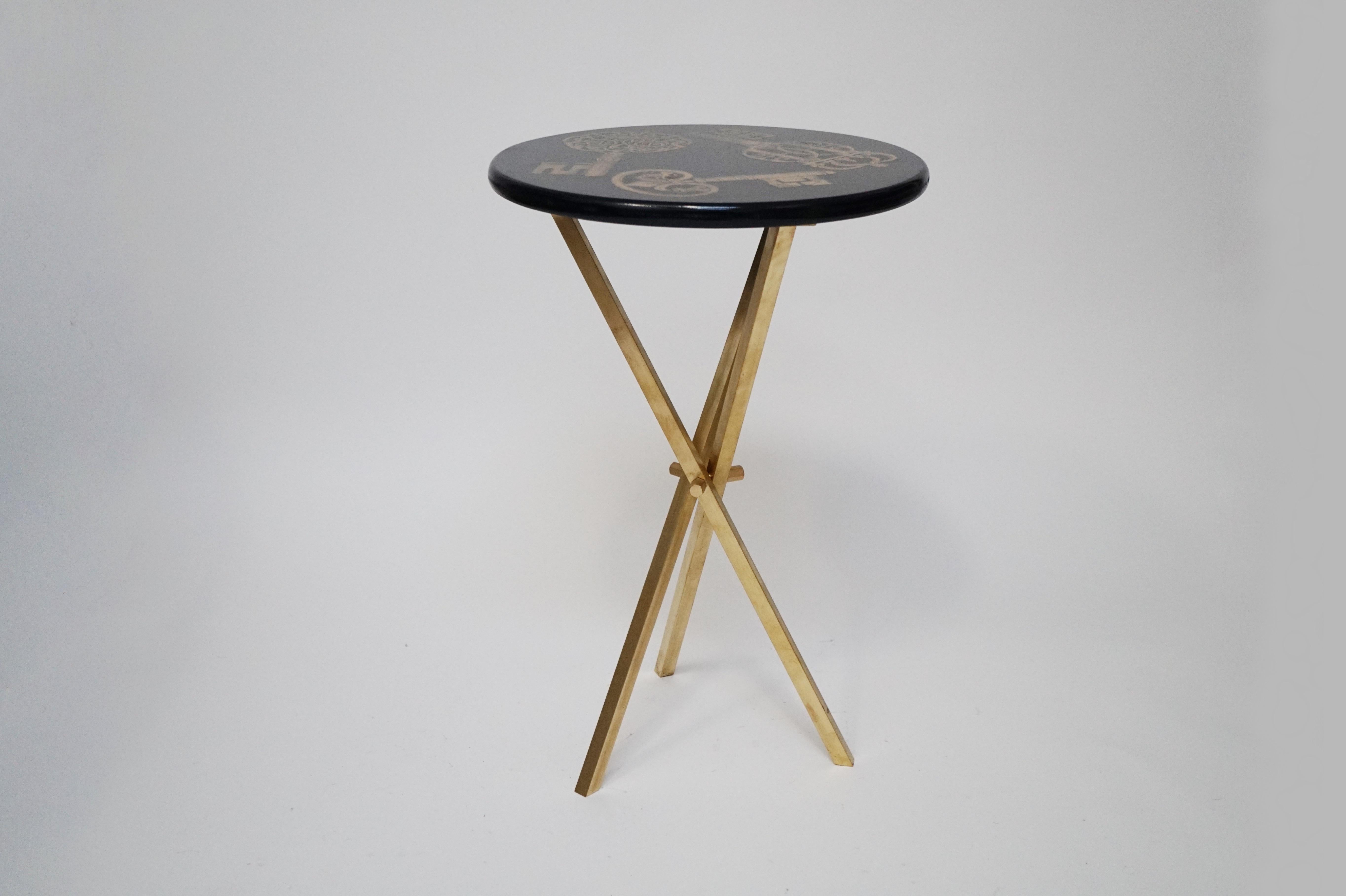 This rare collectors item is the 'Chiavi' side table by Piero Fornasetti, signed underneath with its original label. The side table is made with lacquered wood that has a black and gold 'keys' motif and is affixed on top of a brass tri-pod base that