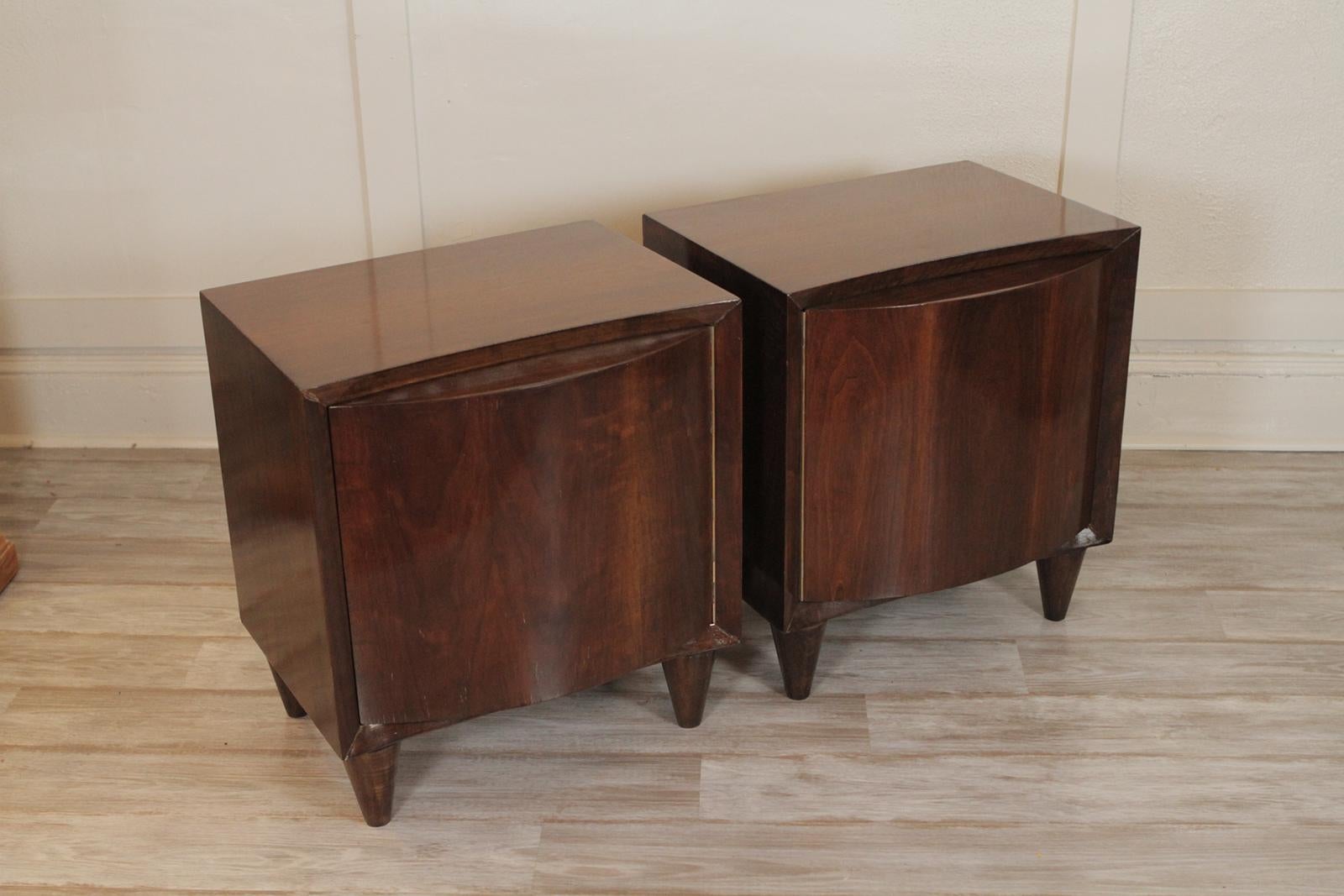 Mid-Century Modern pair of walnut side or end tables. The figurative grained curved door fronts open to reveal a shelved storage area. The outside case in a neat cube form resting of four tapering legs. Dimensions 22” W x 18.5