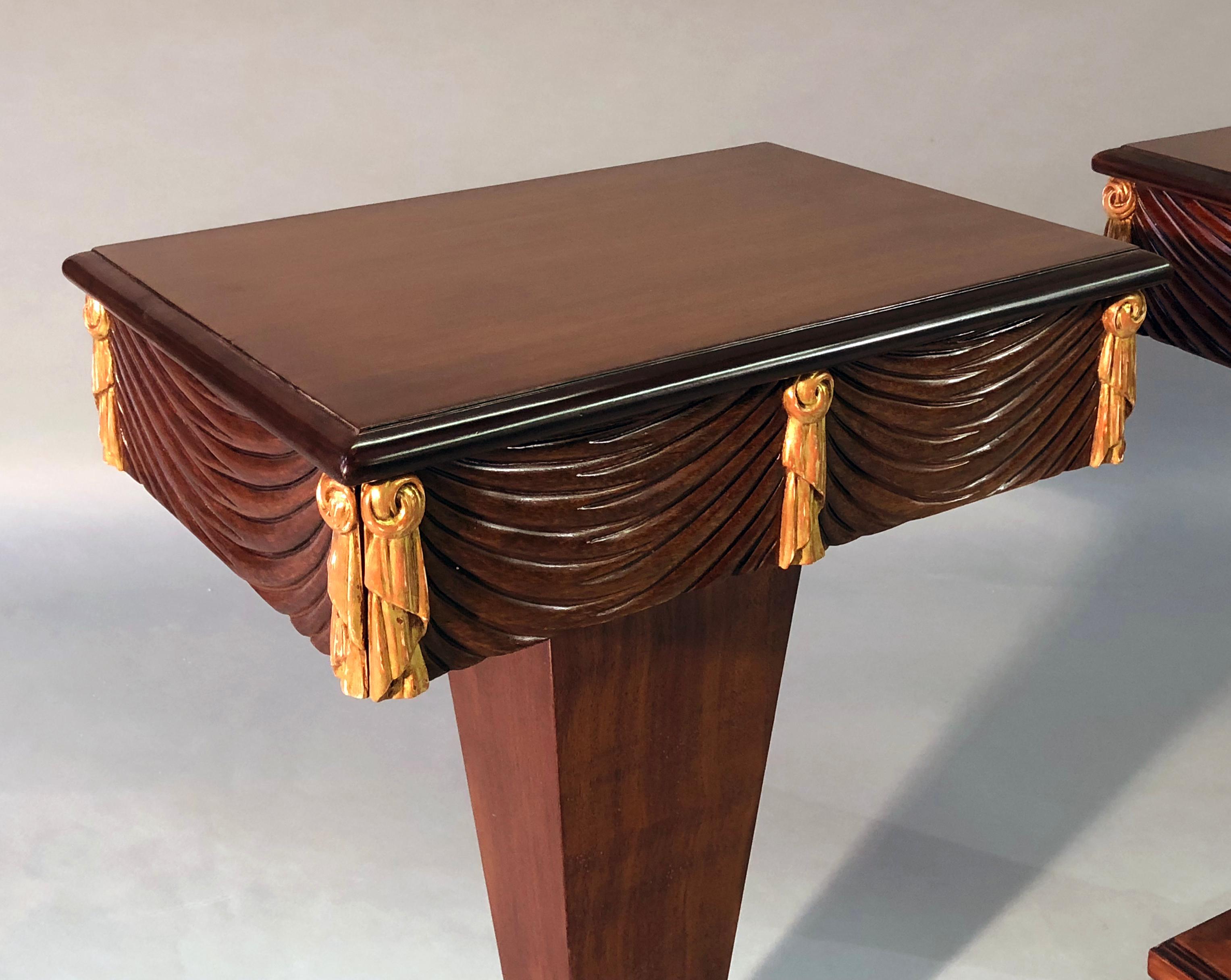 A chic pair of Grosfeld House 1940s mahogany and parcel-gilt single-drawer pedestal tables; each with rectangular top above a swag-carved apron with gilt-wood tassel decoration all fitted with a single drawer; raised on a square tapering supports