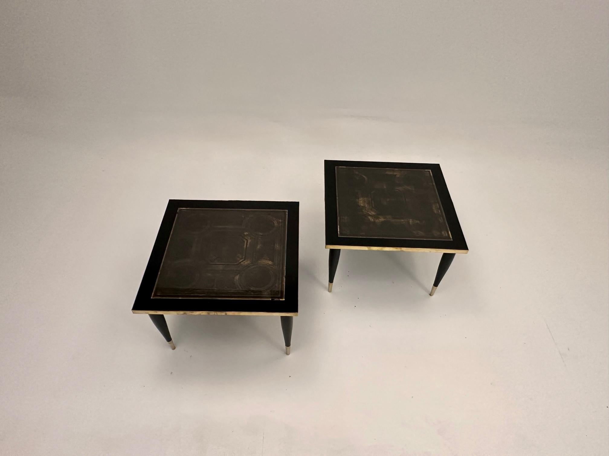 Chic pair of black and gold square low end tables in the style of Fornasetti having reverse painting on glass tops with stylish decorative neoclassical forms.  The legs are tapered and terminate in brass caps.  There's also brass banding around the