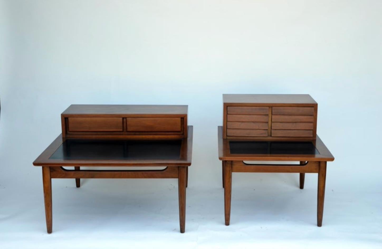 Two chic matching of vintage Mid-Century Modern end tables featuring louvered drawers and a bottom tier with a black laminate top. A sleek two-tier design made of walnut that sits on top of four tapered legs. Quality construction with four sculpted