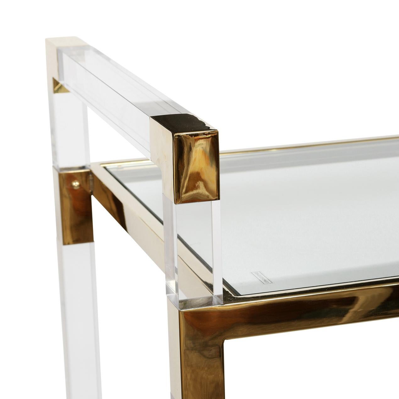 Questo carrello da bar Chic dona un fascino moderno a qualsiasi spazio di intrattenimento. Realizzato in acrilico trasparente, vanta eleganti accenti in ottone e ripiani in vetro temperato, creando un look sofisticato e arioso. Il design trasparente