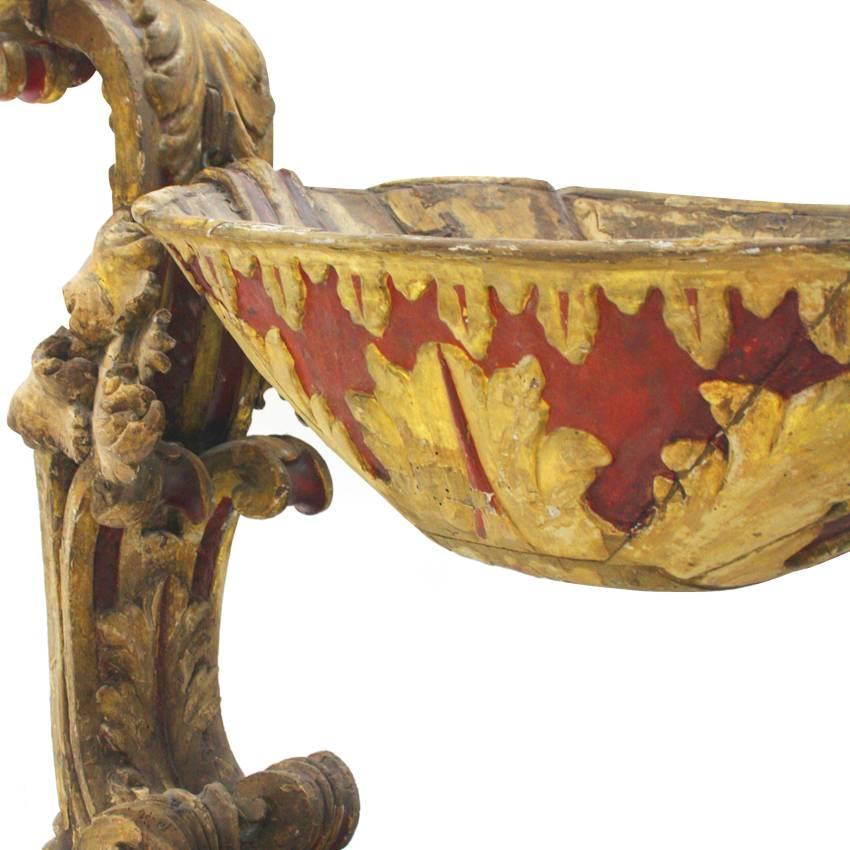 Italian child’s crib standing on a low wooden base with a red and gilt paint. Two carved volute posts support the oval basinet. A female head, wearing an unusual headpiece, marks the front as a figurehead.