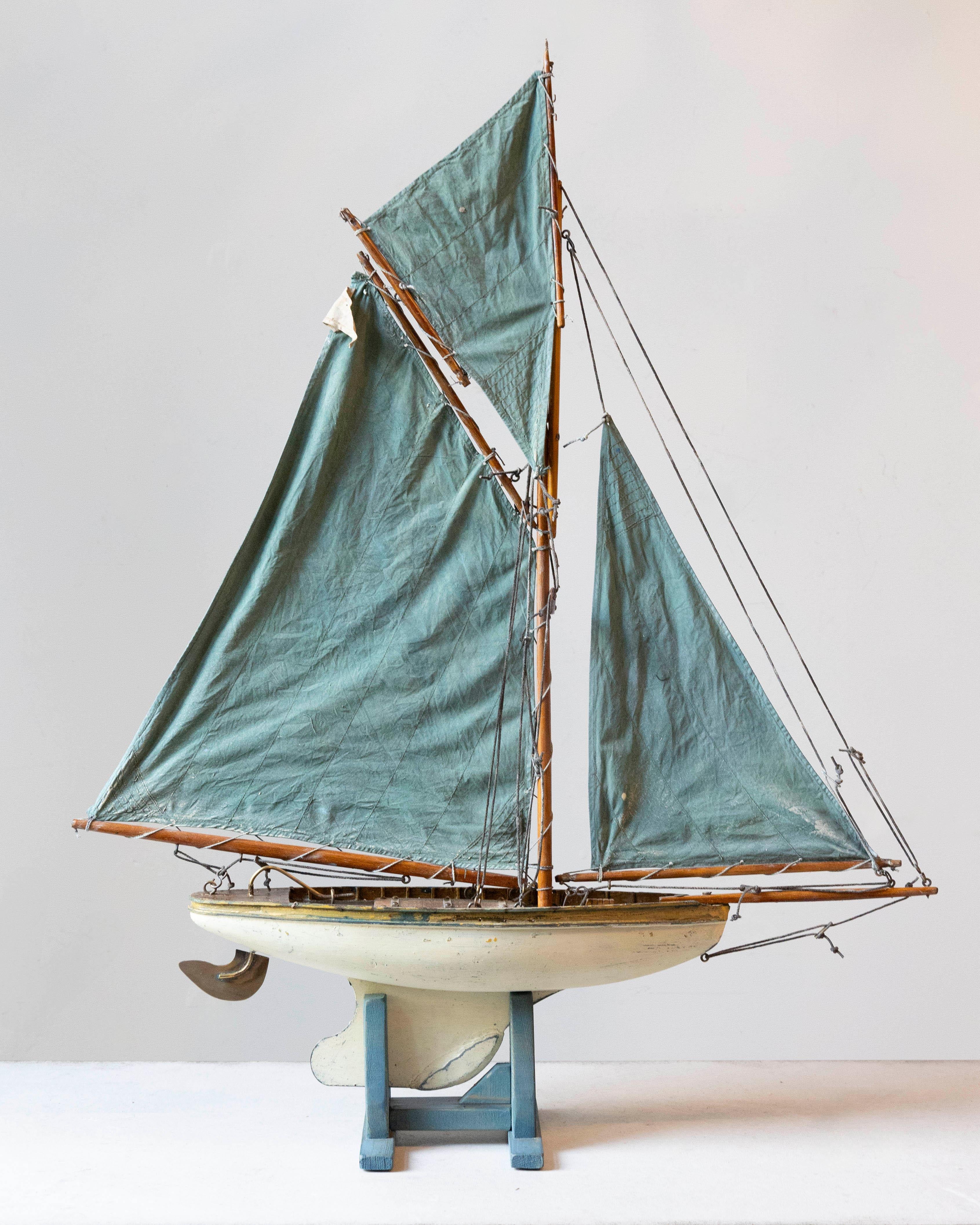 A charming early 20th-century English child’s pond yacht, featuring a rounded white-painted hull with visible age and wear that speak to its playful past and well love history. The yacht is rigged with gaff sails in a striking teal-green cotton,