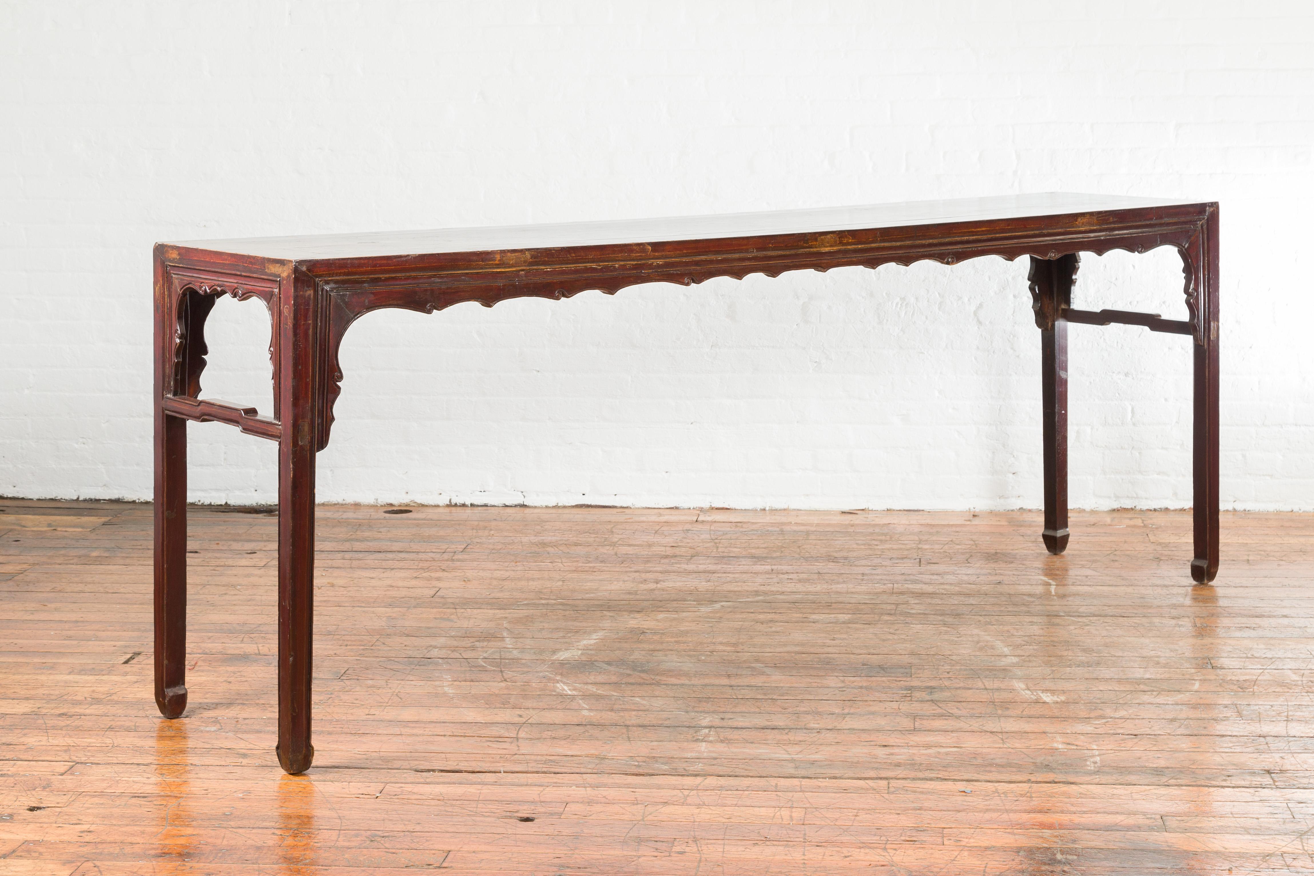 A Chinese Qing Dynasty period altar console table from the 19th century, with dark reddish brown patina. Created in China during the Qing Dynasty, this console table features a rectangular top with central board, sitting above a carved apron with
