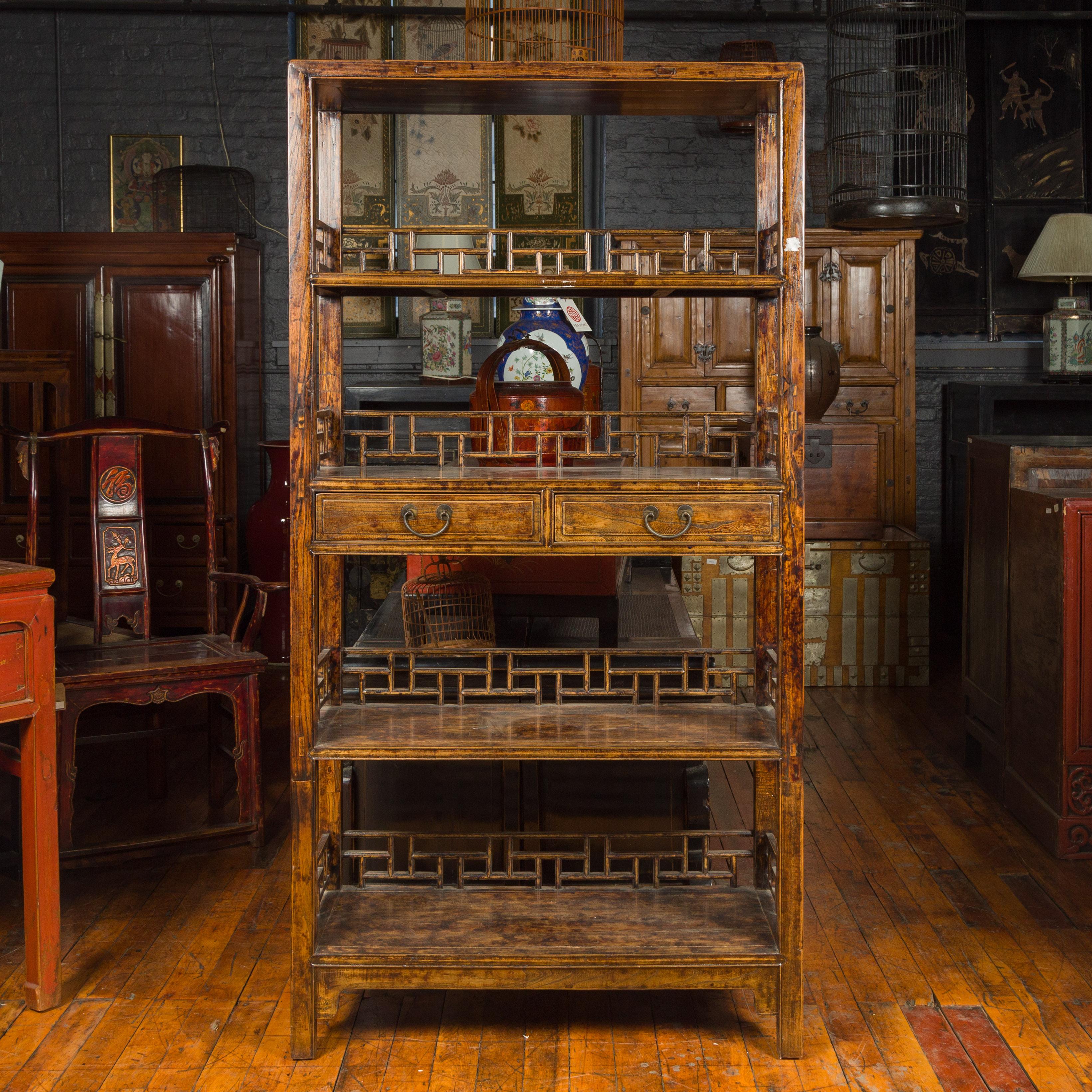 A Chinese Qing Dynasty period stained elm bookcase from the 19th century, with fretwork sides, open shelves and two drawers. Crafted in China during the 19th century, this elm bookcase features a linear silhouette presenting open shelves, each