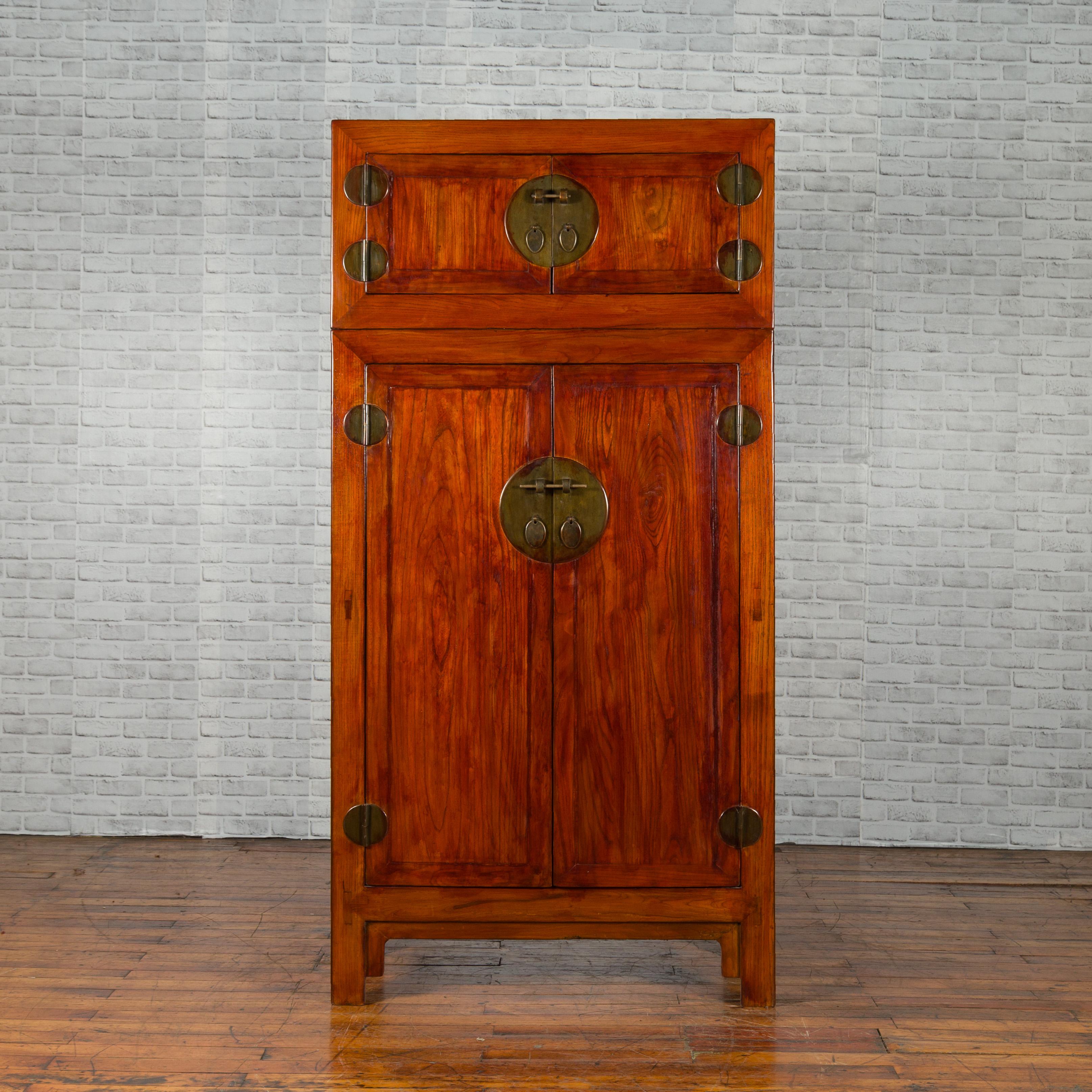 A Chinese Qing Dynasty period compound elm cabinet from the 19th century, with reddish brown lacquer and traditional brass hardware. Created in China during the Qing Dynasty, this compound two-piece cabinet features a linear silhouette perfectly