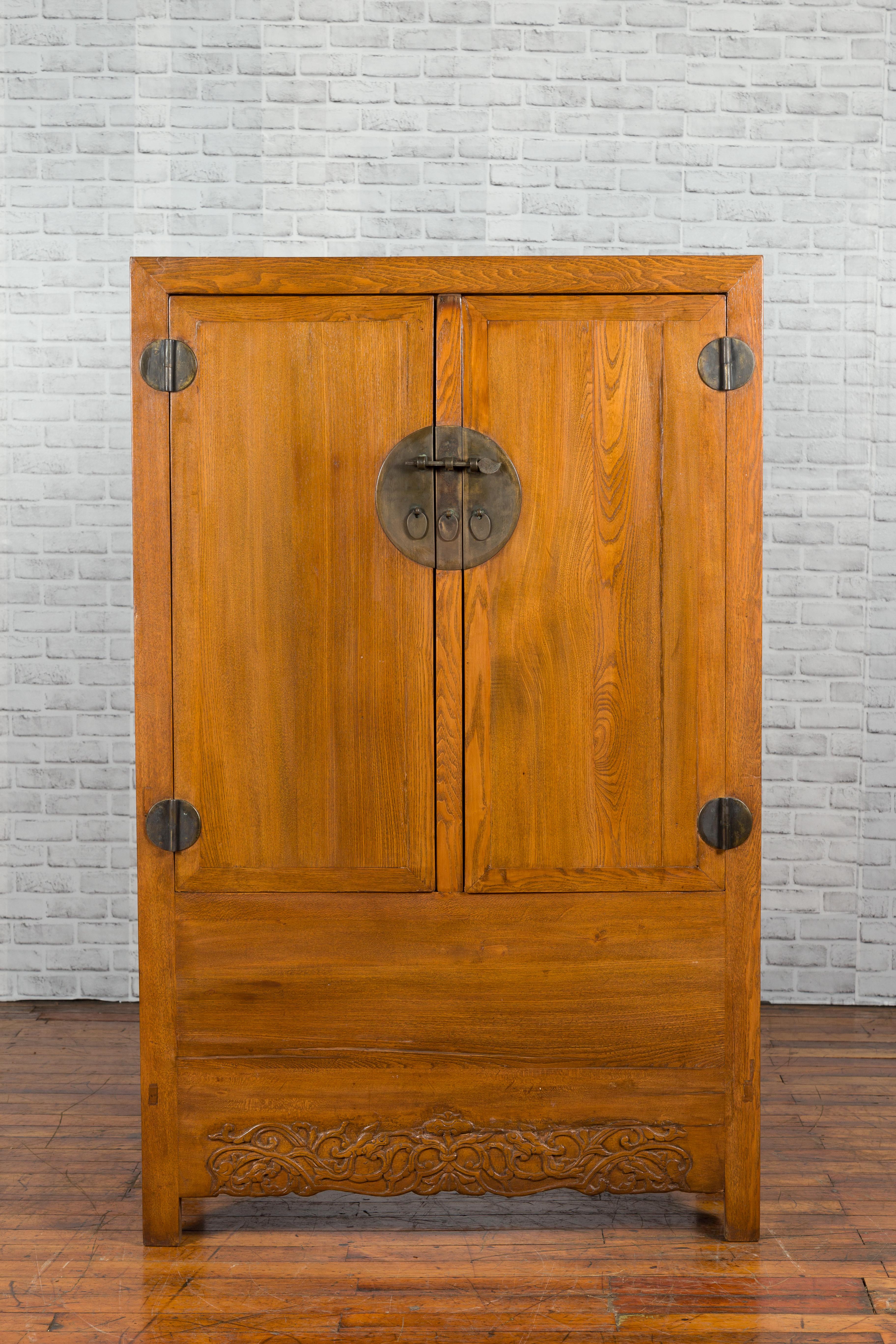 A Chinese Qing dynasty period natural elm cabinet from the 19th century, with carved apron and brass medallion. Created in China during the Qing dynasty, this cabinet features a linear silhouette perfectly complimented by its natural elm finish. Two