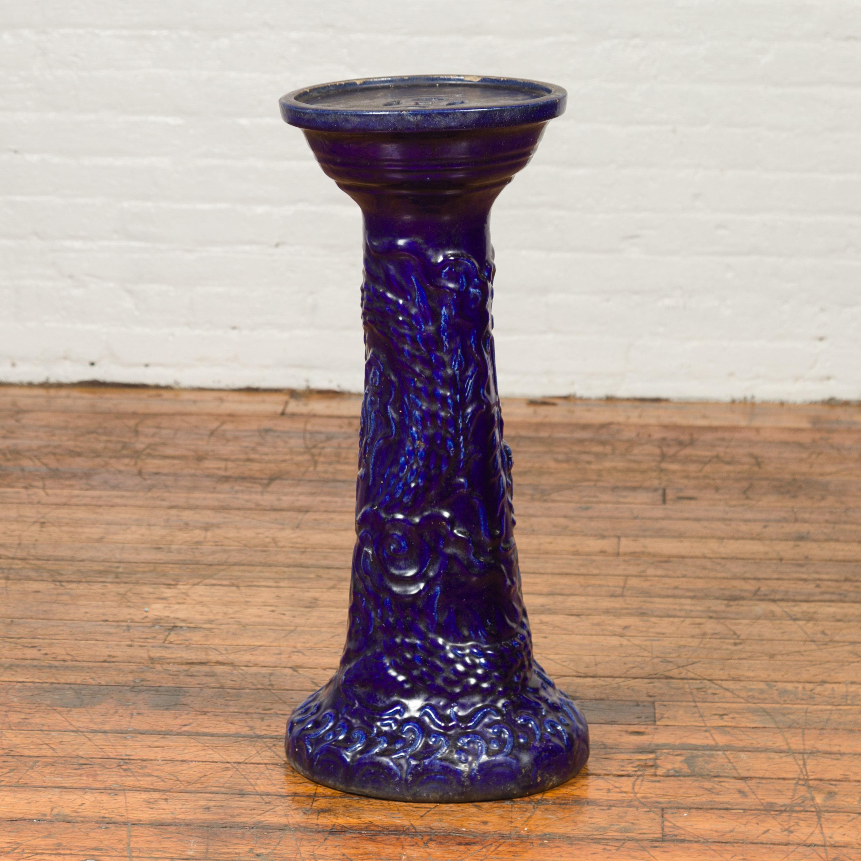 A Chinese antique dark blue glazed artisan pedestal stand with scrolling effects. Boasting a lovely dark blue glazed patina, this plant stand features a circular top pierced with small geometric openings, resting above a pedestal discreetly accented