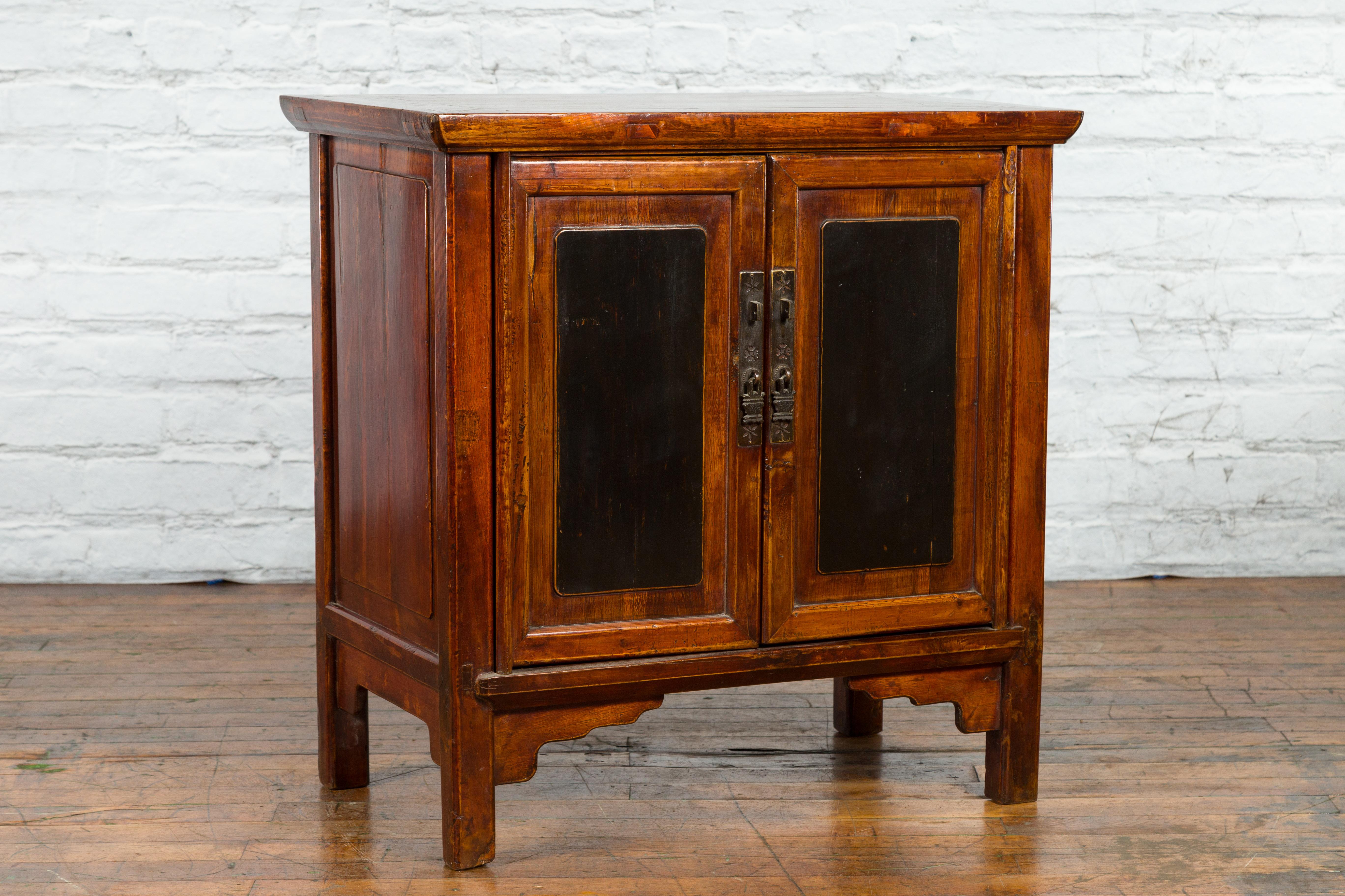 A Chinese antique side cabinet from the early 20th century with brown and black lacquer, brass hardware and carved spandrels. Created in China during the early years of the 20th century, this side cabinet features a rectangular top with central