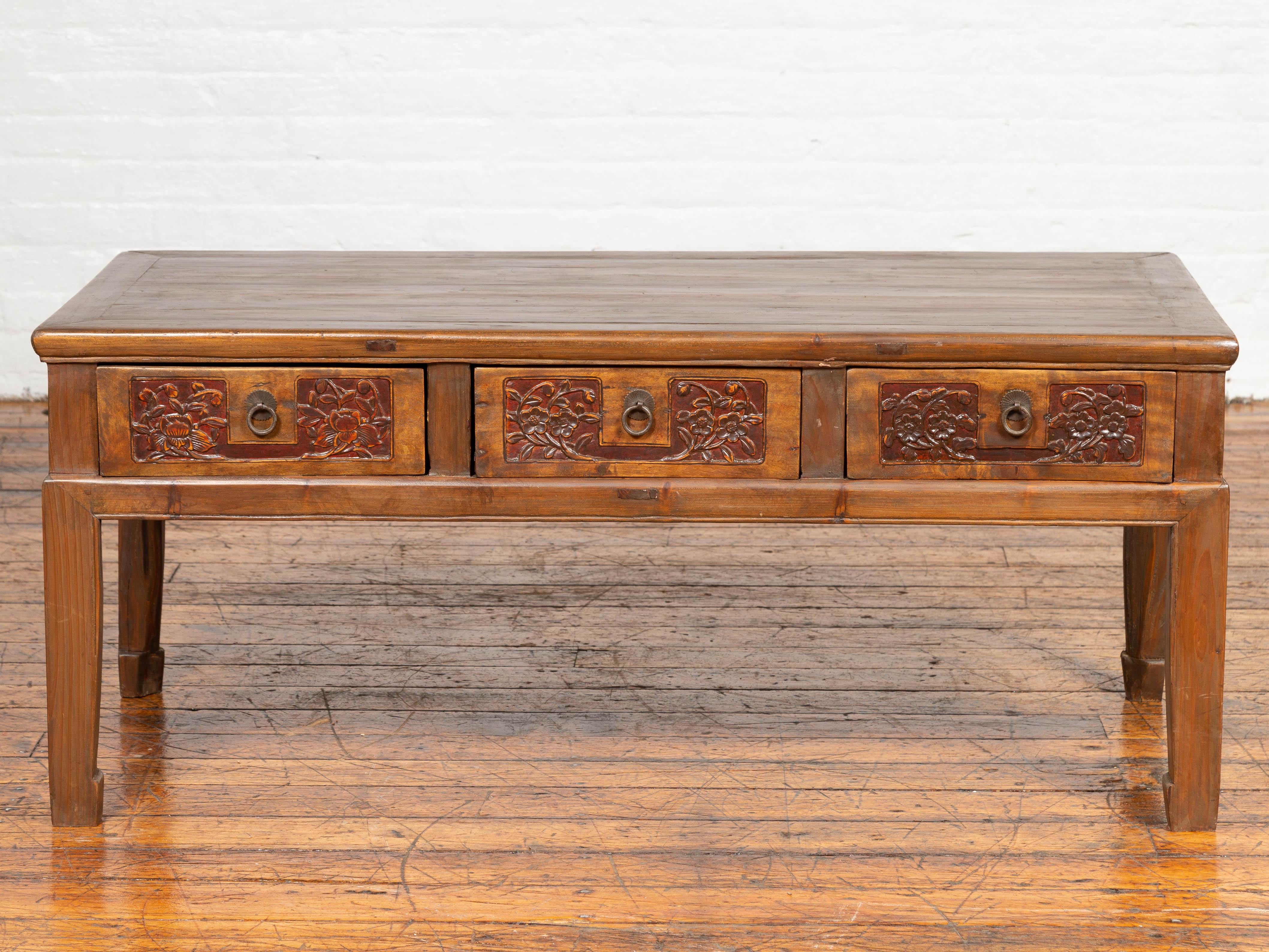 An antique Chinese elmwood coffee table with three carved drawers and horsehoof feet. Featuring a rectangular top with central board, this Chinese coffee table presents three drawers, each adorned with floral motifs carved in low-relief, standing
