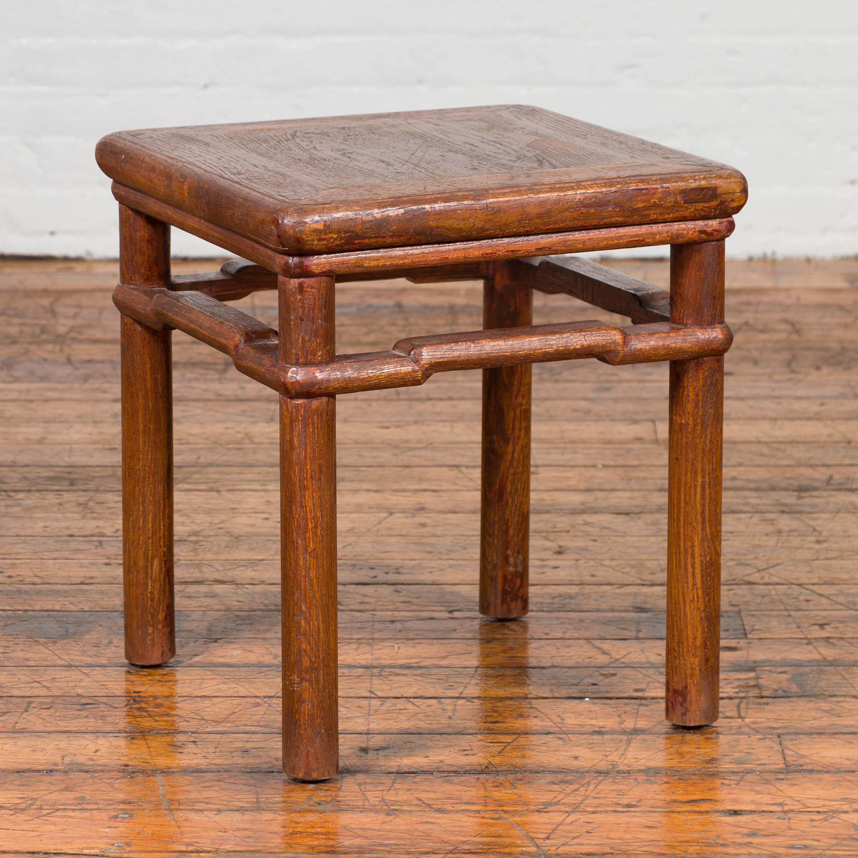 An antique Chinese Ming dynasty style distressed wood side table from the early 20th century, with humpbacked stretcher and cylindrical legs. Originally conceived as a stool, this side table features a square top with central board and rounded