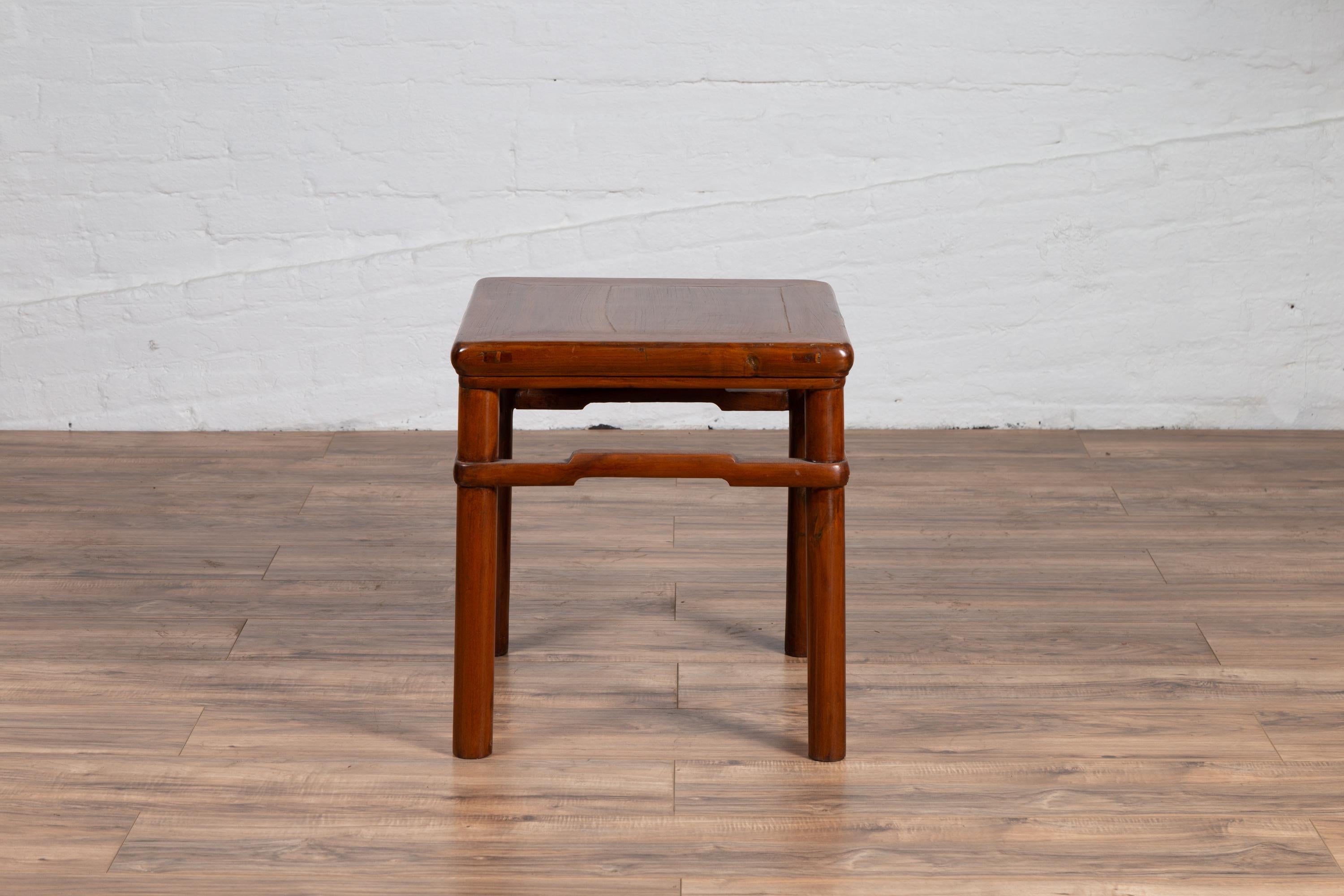 An antique Chinese Ming dynasty style wooden side table from the early 20th century, with humpbacked stretcher and cylindrical legs. Originally conceived as a stool in China where it was born during the early years of the 20th century, this charming