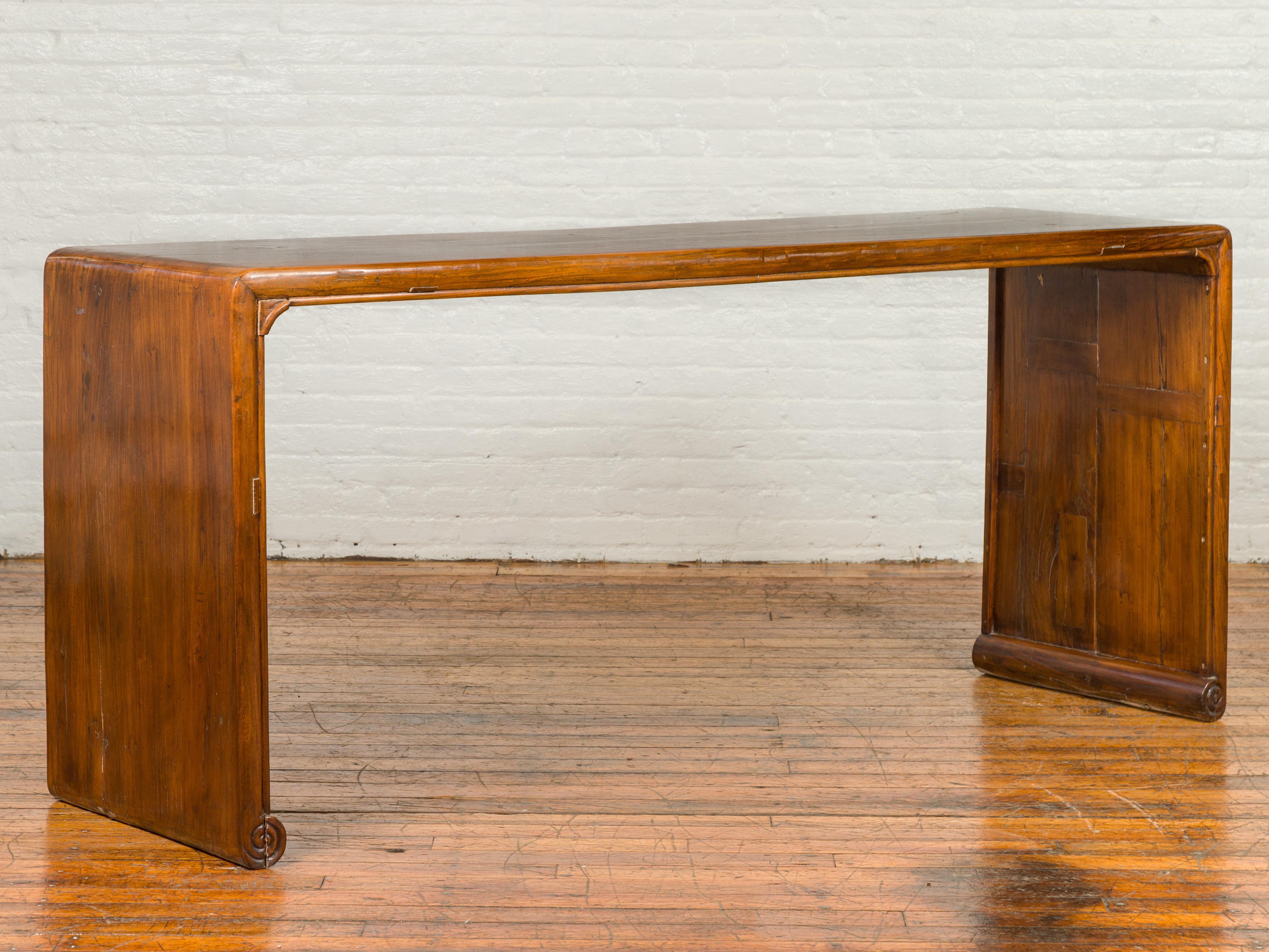 An antique Chinese Qing Dynasty waterfall console table from the 19th century, with scrolling feet and warm patina. Crafted in China during the 19th century this console table features a waterfall wooden frame with solid boards, boasting a lovely