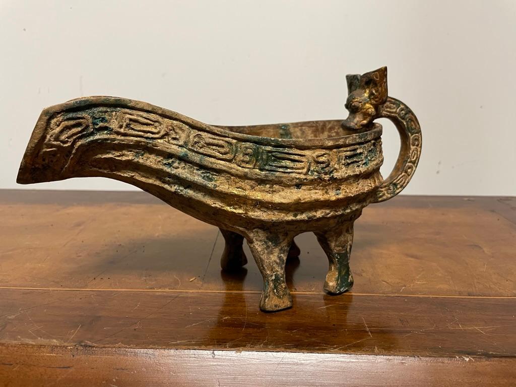 Vasija vertedera china de bronce dorado, de estilo arcaico, con asa en forma de cabeza de dragón y decoración geométrica. Con una maravillosa pátina dorada y verdigris. Recipiente en forma de barco con asa de un solo lazo apoyada en cuatro patas.