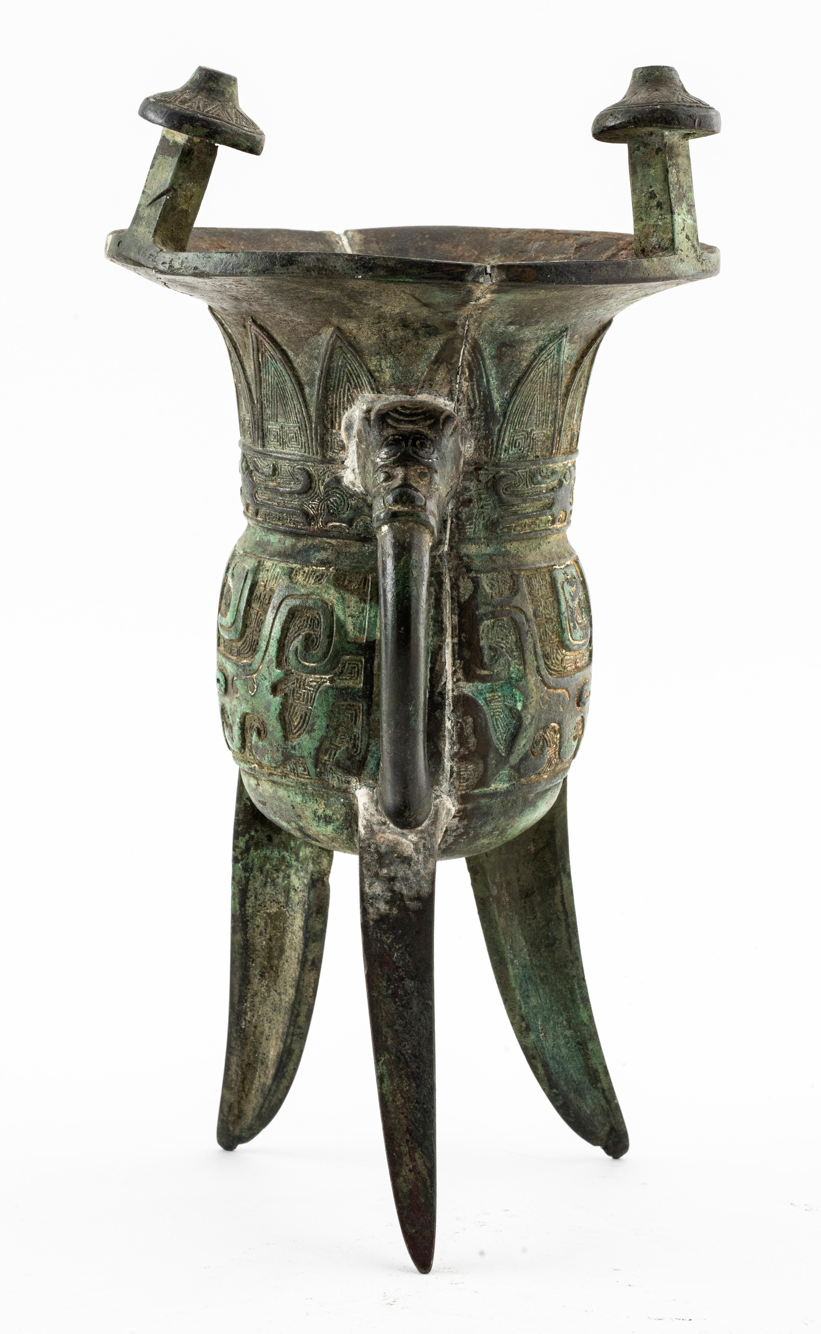 Chinese archaic style jue bronze libation vessel. The bublous body raised on tripod blade-shaped supports and cast in high relief with a pair of ta'ot'ien masks on a fretwork ground below the wide trumpet neck, with erect leaf-tip motifs, a loop