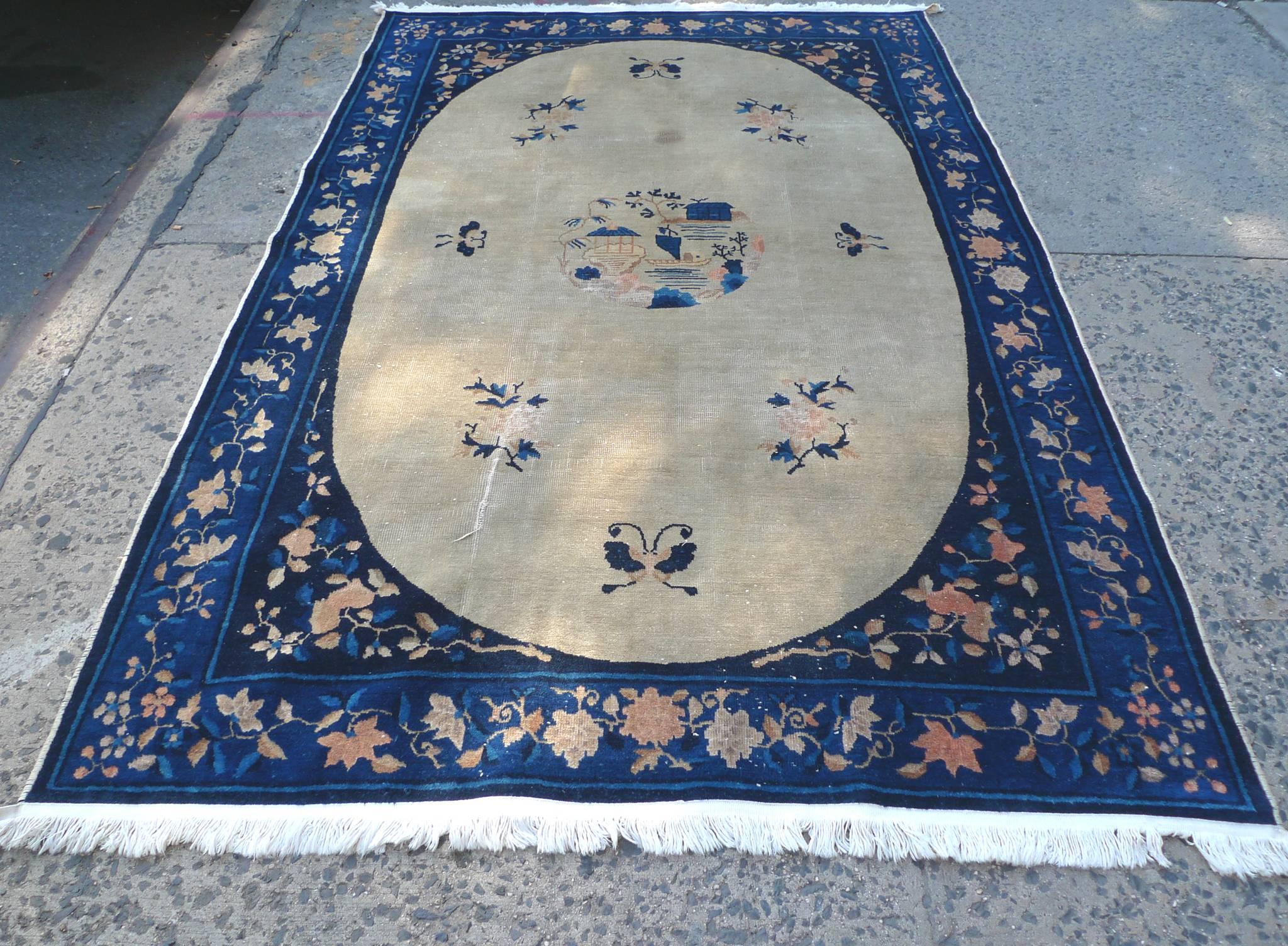 This Chinese Art Deco rug was manufactured by Walter Nichols, circa 1930s-1940s. Its designs consists of a central white oval within a rich blue rectangle, which is in turn surrounded by a blue border. Floral patterns in pink hues adorn the border