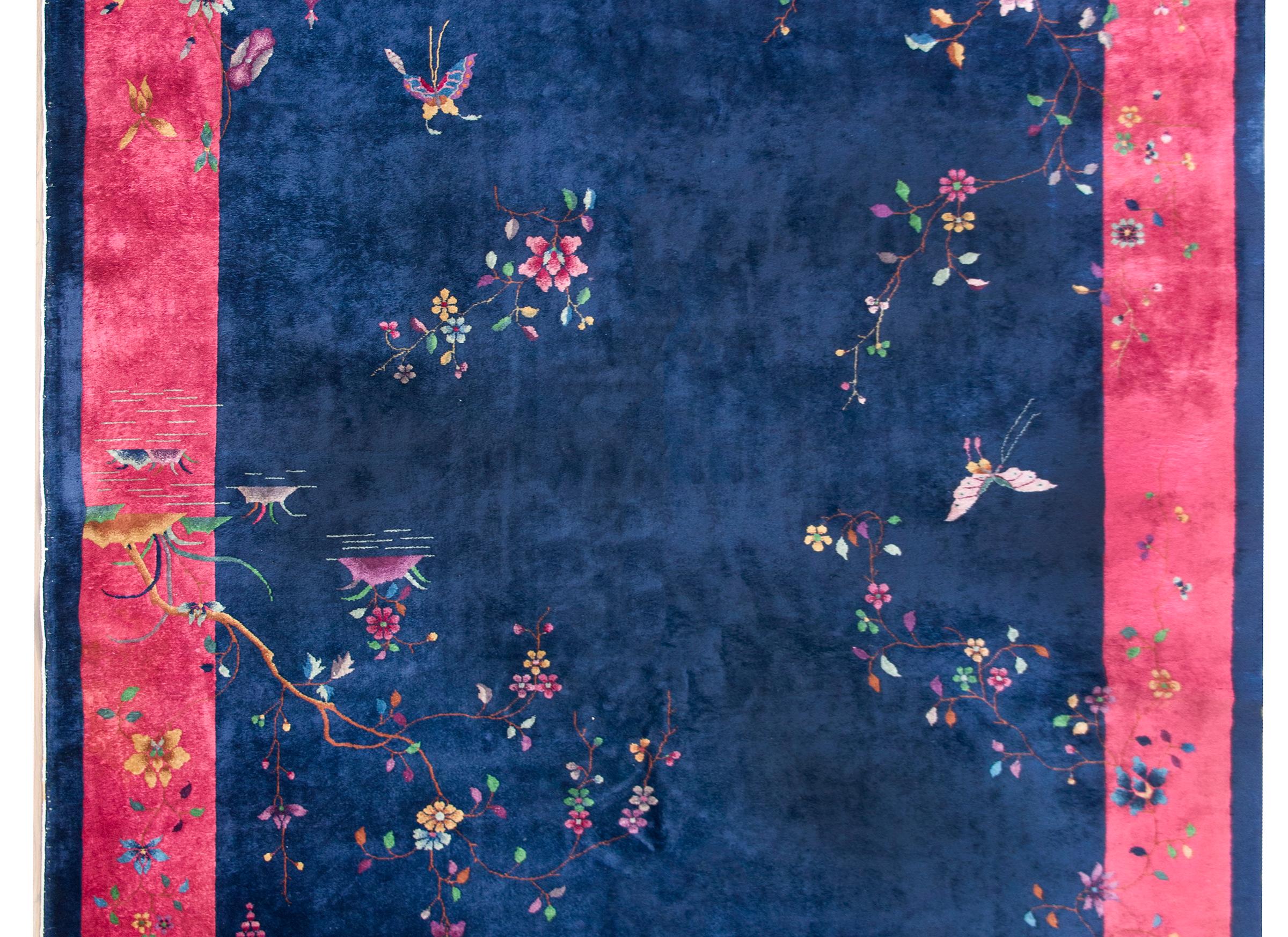 A beautiful early 20th century Chinese Art Deco rug with a dark indigo background surrounded by a wide fuchsia border, and overlaid with wispy prunus blossoms and peonies, butterflies, and a garden pagoda in the lower right corner.