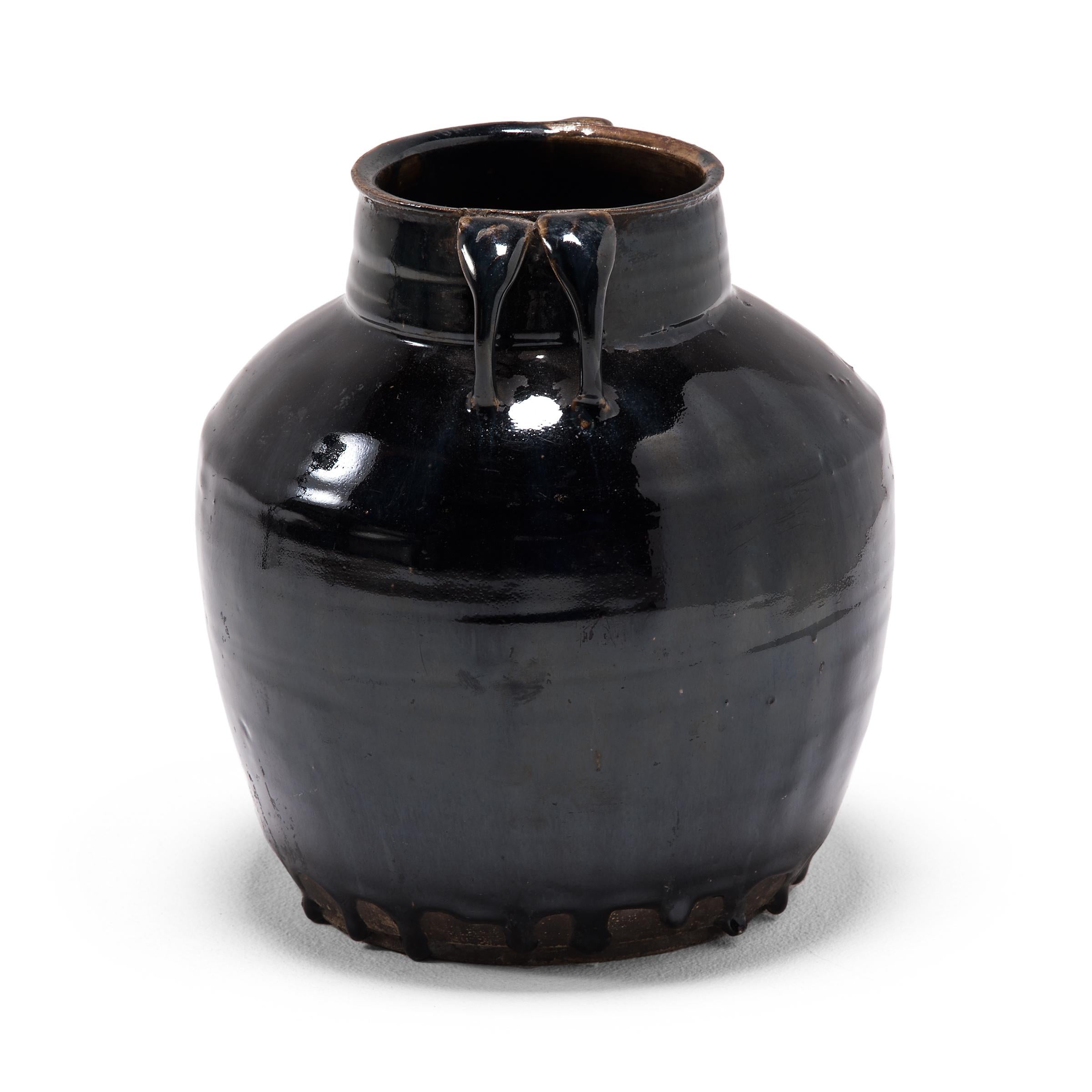 As though slicked with oil, an inky black glaze drenches the high shoulders and arched double strap handles of this tapered kitchen vessel. The 19th century ceramic jar was once used to prepare and store soy sauce in a Qing-dynasty kitchen, as