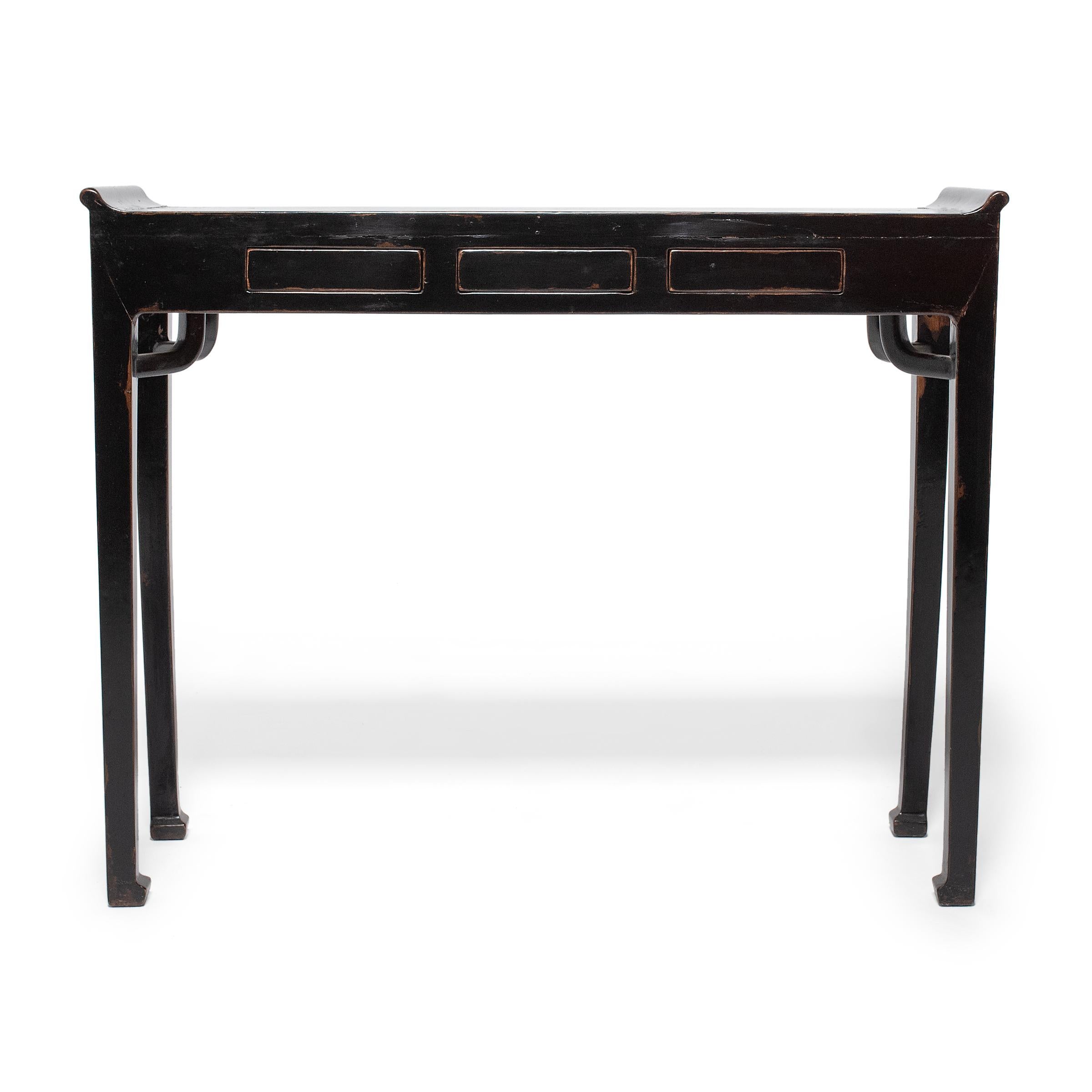 This black lacquer console table dates to the early 20th century and has the narrow form and everted flanges of a traditional altar table. The table has a flush-sided corner leg design with straight legs ending in hoof feet and topped by elegantly