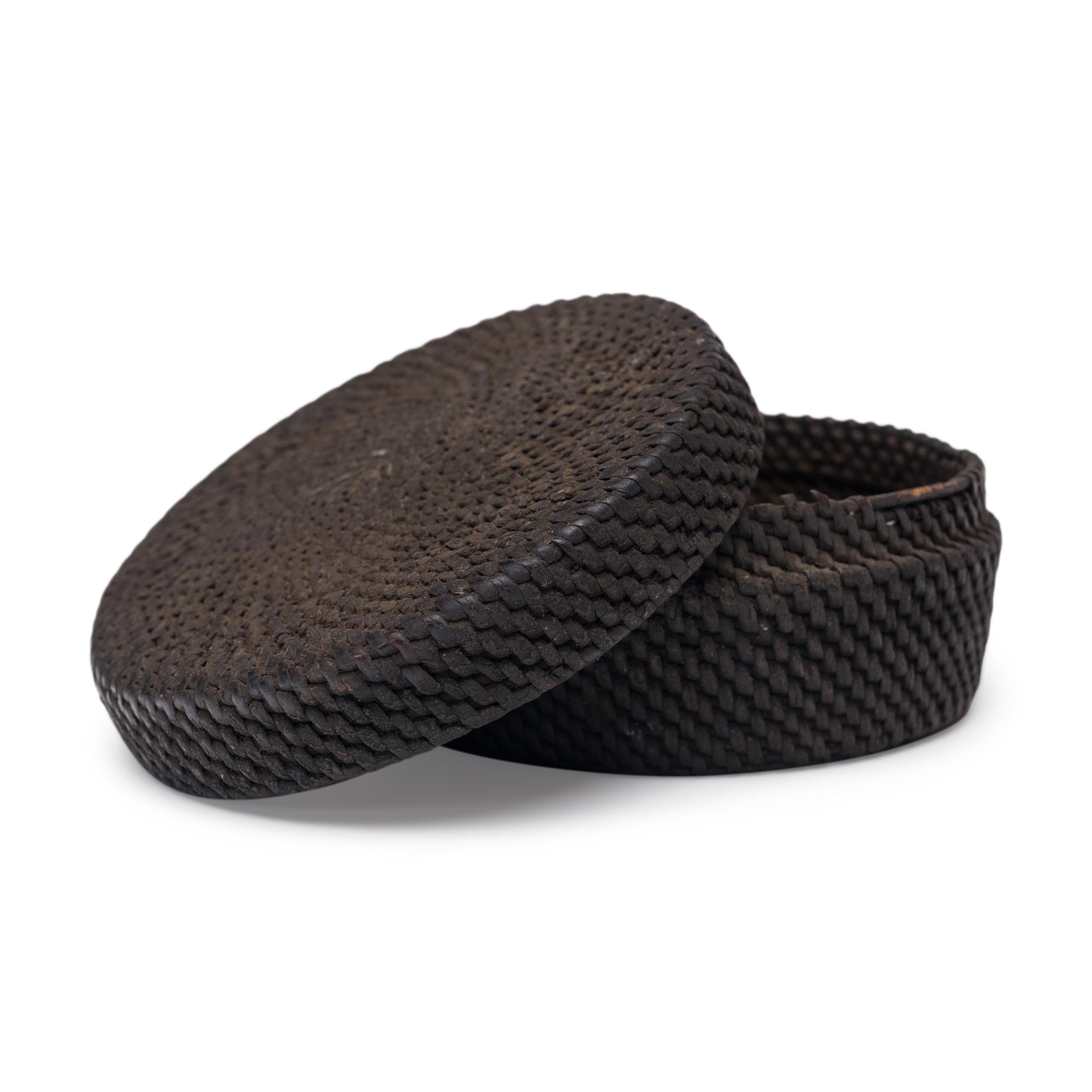 This small woven box from the early 20th century celebrates the beautiful simplicity of a hand-woven object. The round, lidded box is shaped from tightly woven reeds and finished with a thick coating of black lacquer. A charming example of a