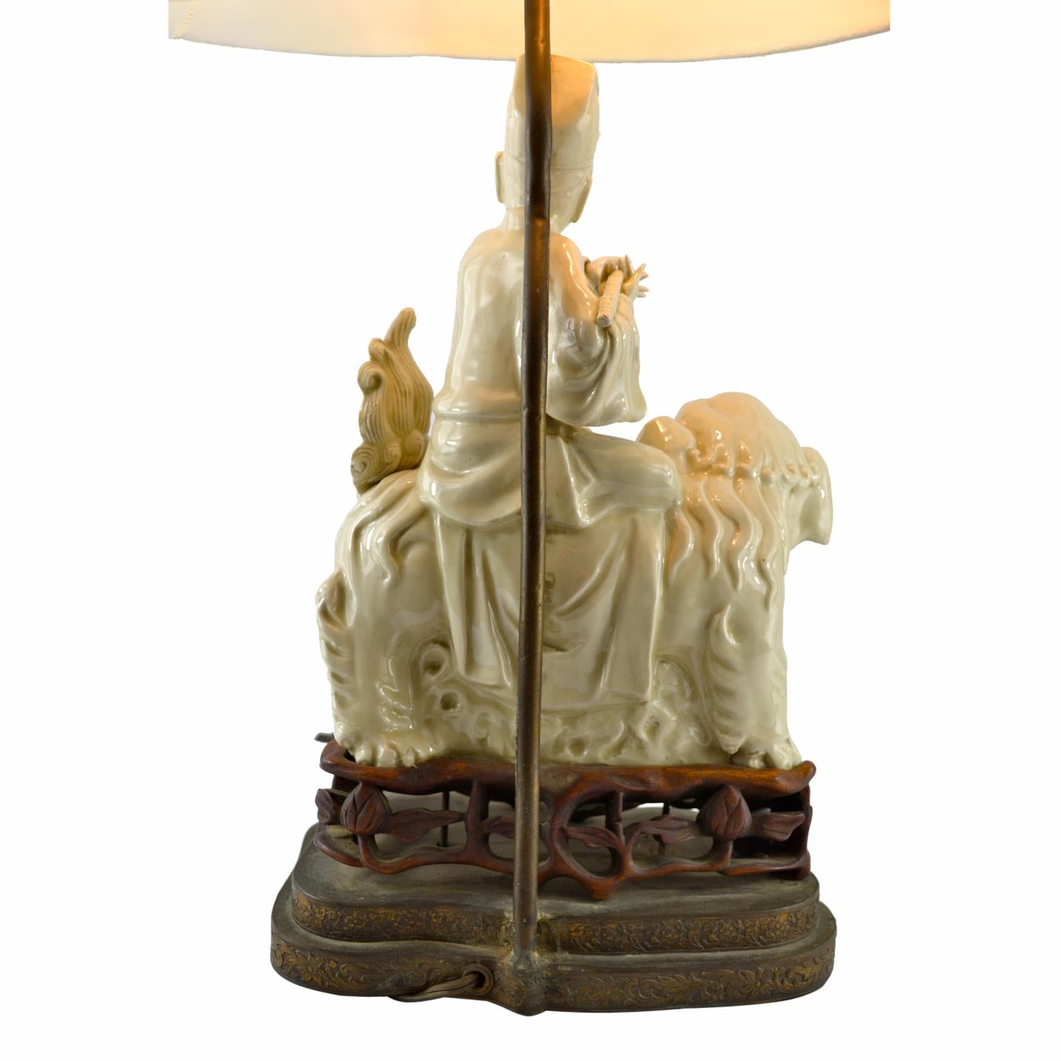A Blanc de Chine lamp featuring a Chinese god sits astride a growling foo dog, the whole mounted on a carved rosewood base on a further gilt metal base.