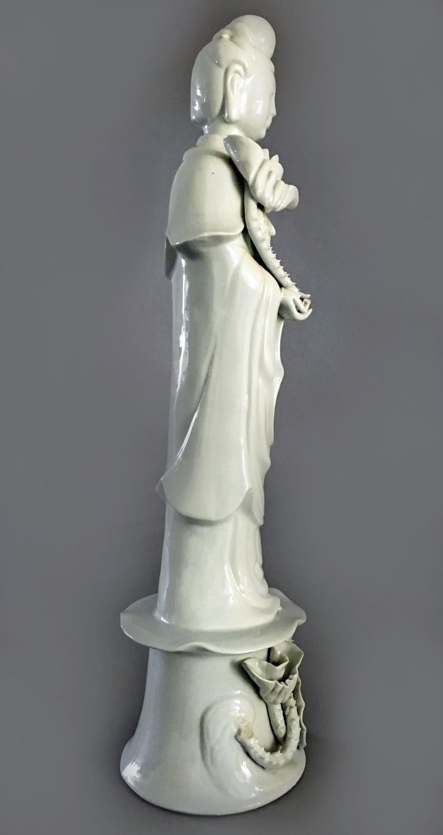 Chinese tall blanc de chine standing Quan Yin dressed in a flowing robe holding a lotus blossom which suggests purity. The base is decorated with lotus flowers and pads.